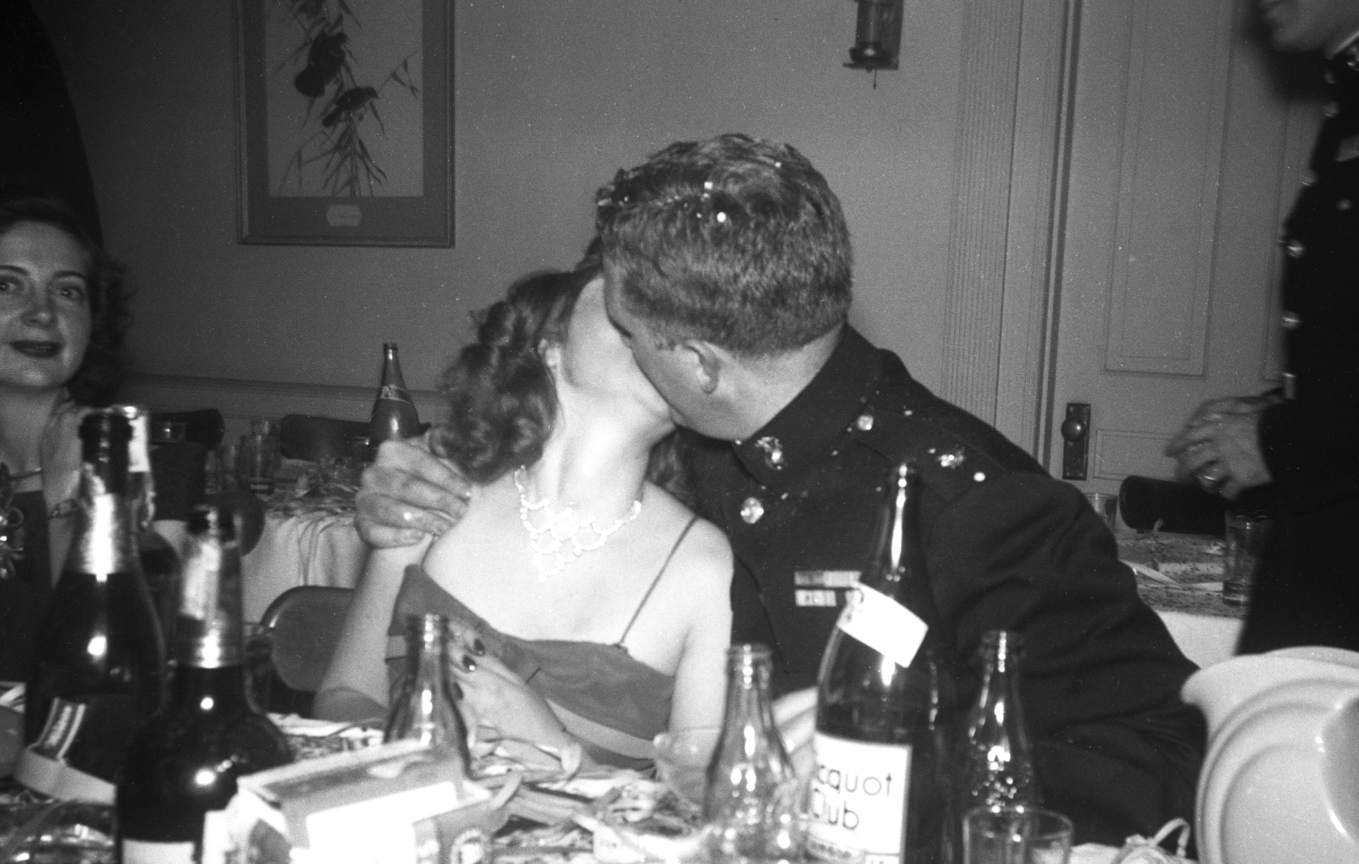 A man in a military uniform kisses a woman wearing a necklace at a dinner table with bottles of alcohol, while a woman to the left watches.