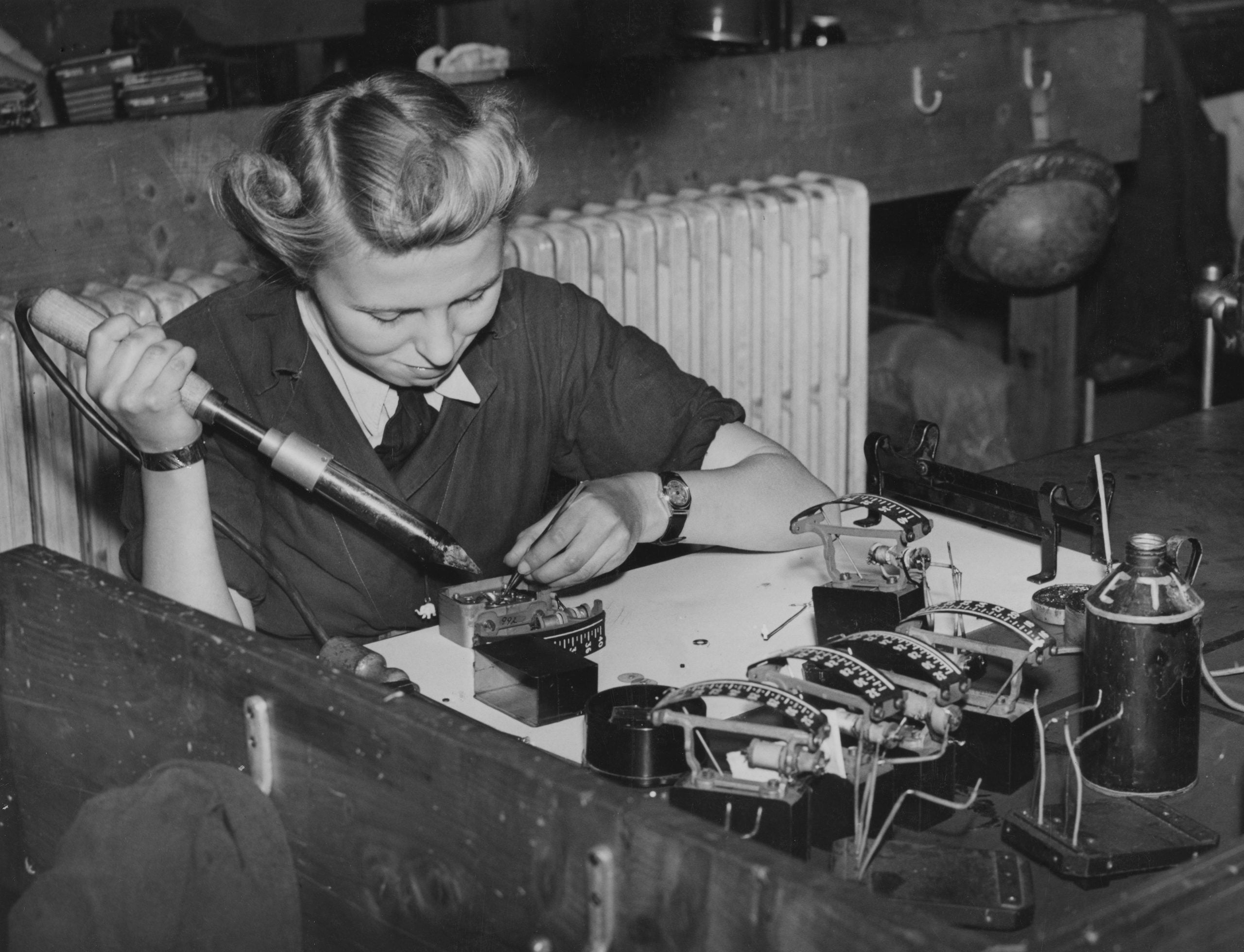 A woman sits at a workbench using a soldering iron to assemble electrical components, with tools and parts scattered around her.