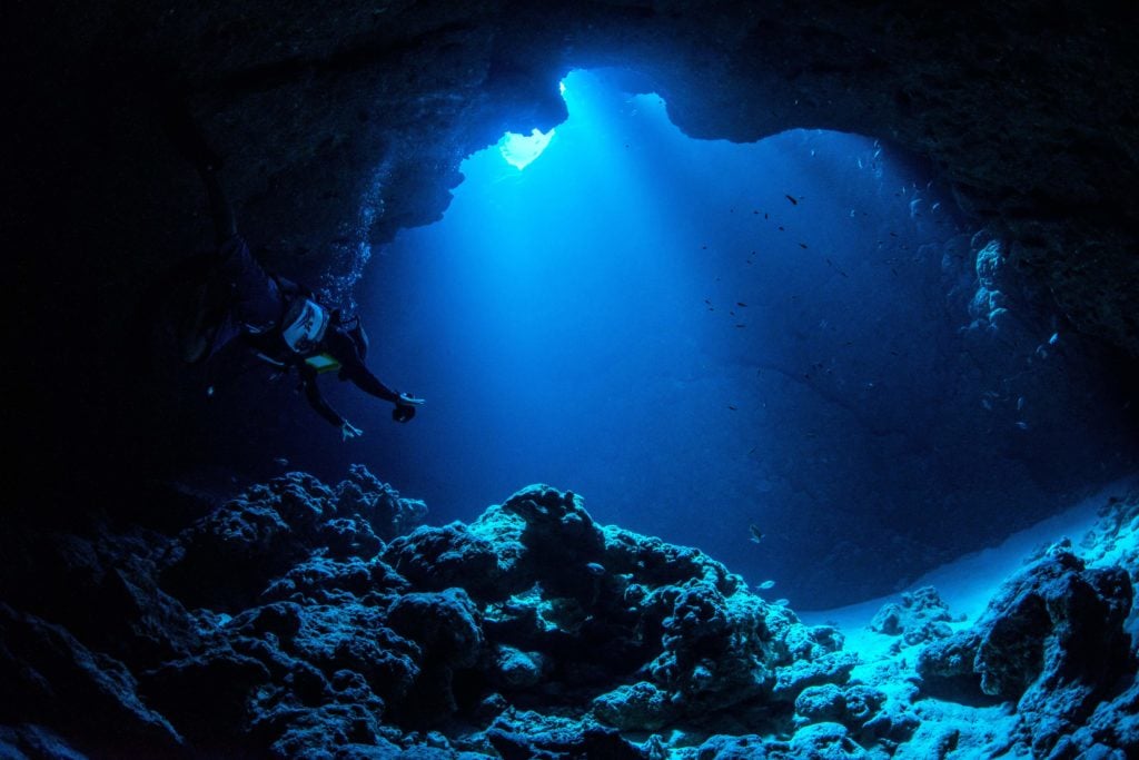 A scuba diver explores a dark underwater cavern lit by a beam of sunlight from above.