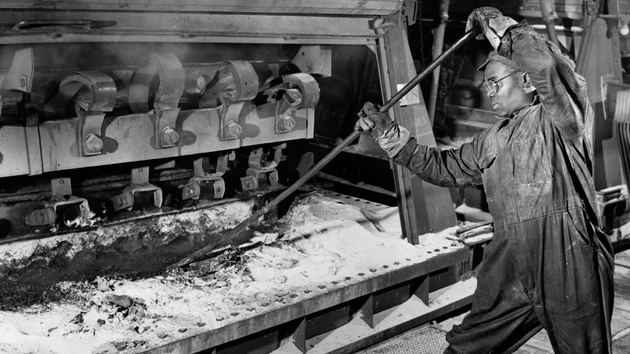 A worker in protective clothing uses a long tool to tend molten material inside a large industrial furnace, with mechanical parts and ash visible.