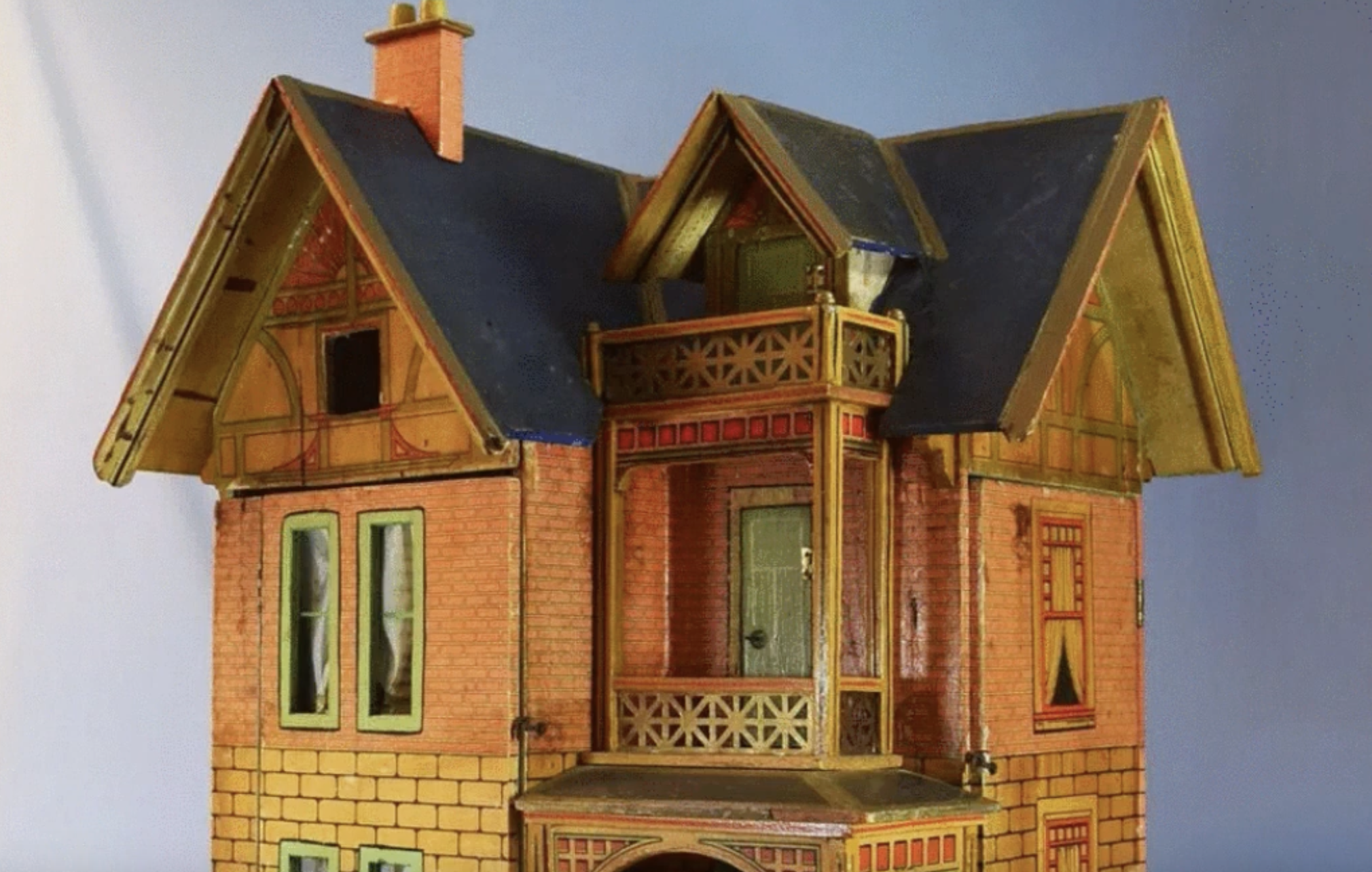 A vintage toy dollhouse with a dark roof, brick-patterned walls, a second-story balcony, and a prominent gable.