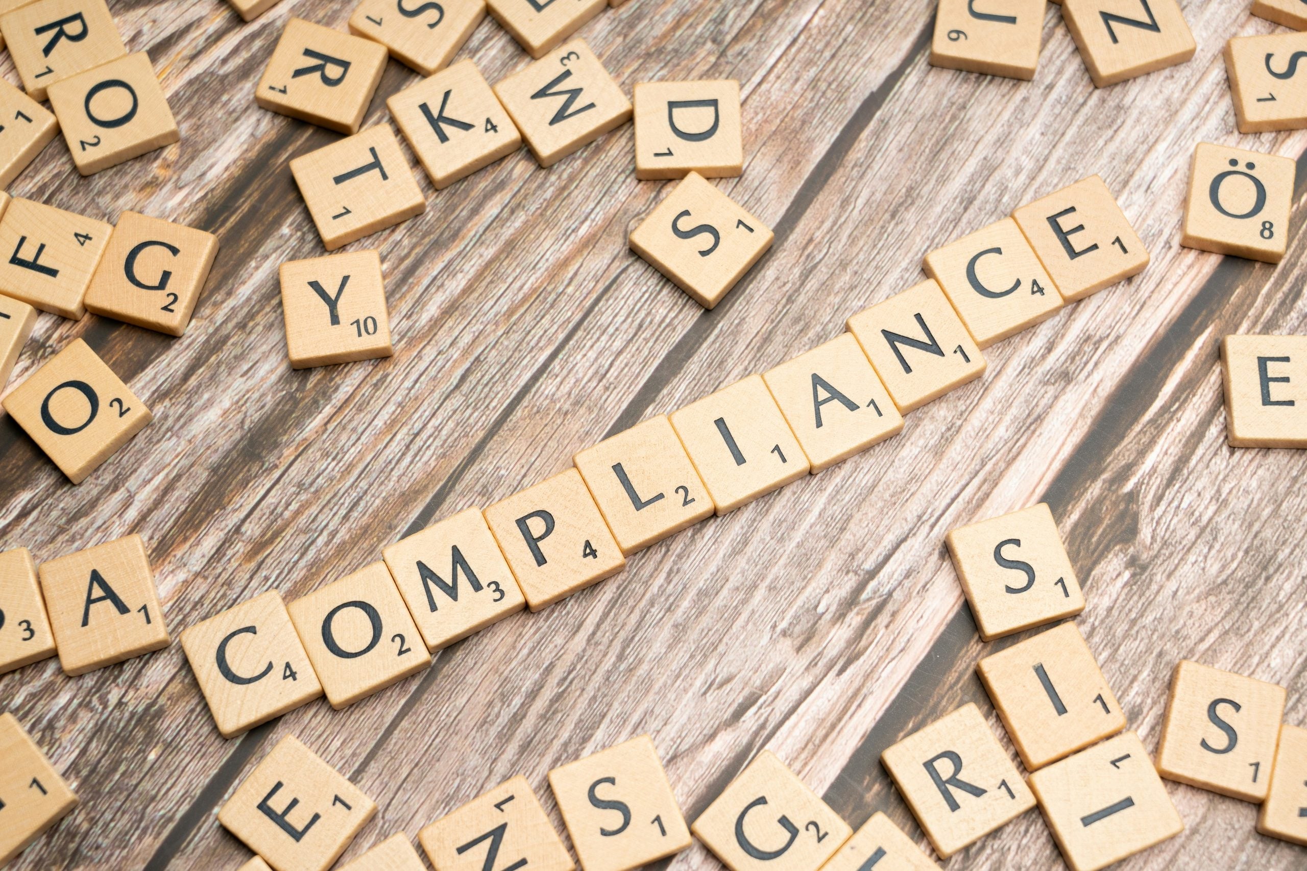 The word “COMPLIANCE” spelled out with wooden letter tiles on a wooden surface, surrounded by scattered loose tiles.
