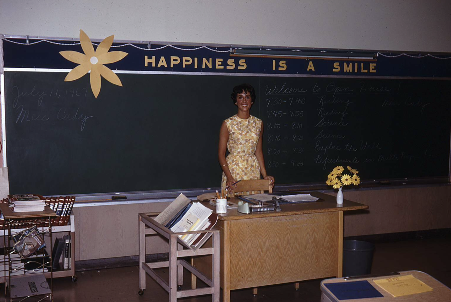 A teacher stands in front of a chalkboard decorated with a large paper flower and the text “Happiness is a smile,” with an Open House schedule written on the board behind her.