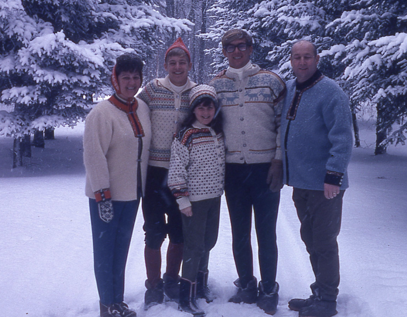 A family of five posing in the snow, wearing patterned sweaters and winter gear among snow-covered pine trees.
