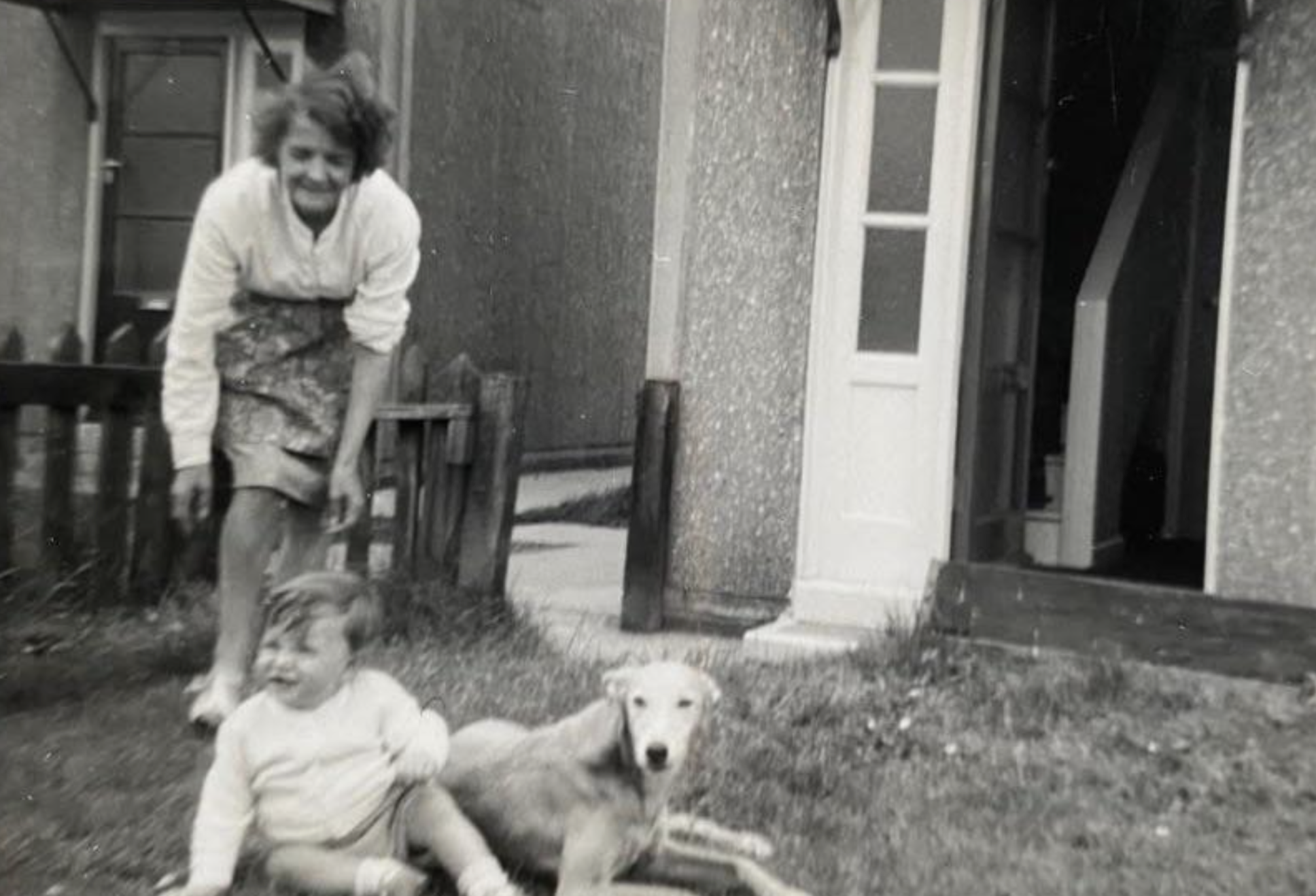 A woman stands in a yard next to a toddler sitting on the grass beside a dog.
