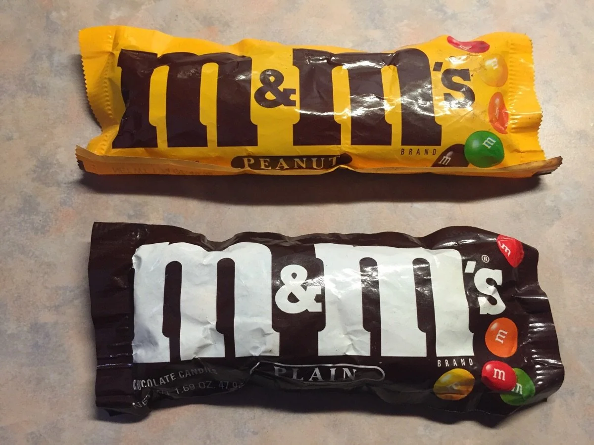 A yellow bag of M&M’s Peanut and a brown bag of M&M’s Plain chocolate candies.