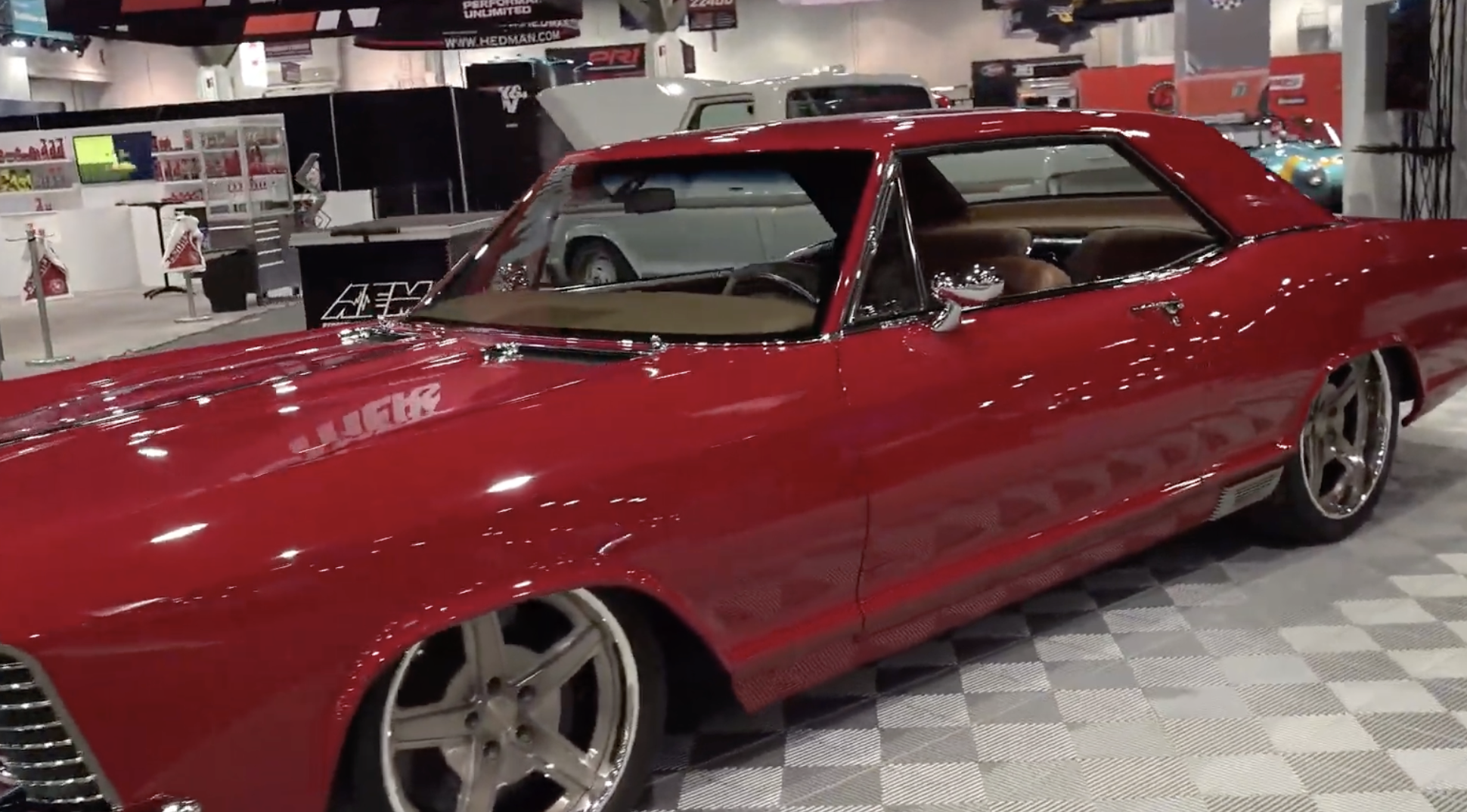 A shiny red classic muscle car with large silver rims is displayed indoors at an auto show. Other cars and display booths are visible in the background.