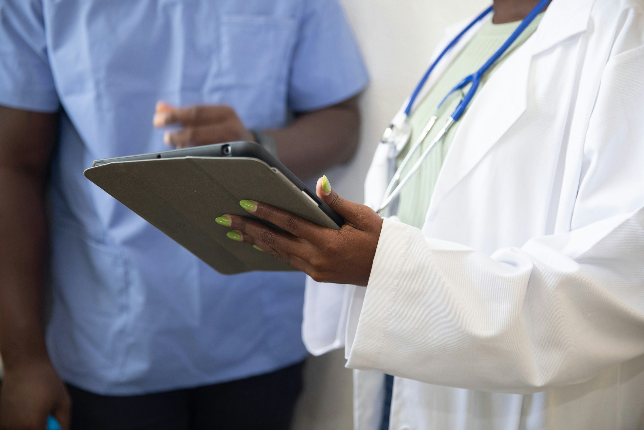 A healthcare professional in a white coat with a stethoscope holds a tablet while consulting with a colleague in blue scrubs.