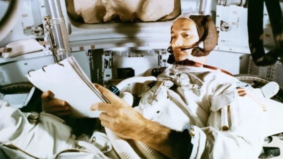 An astronaut in a spacesuit sits inside a spacecraft cabin, looking down at a clipboard.