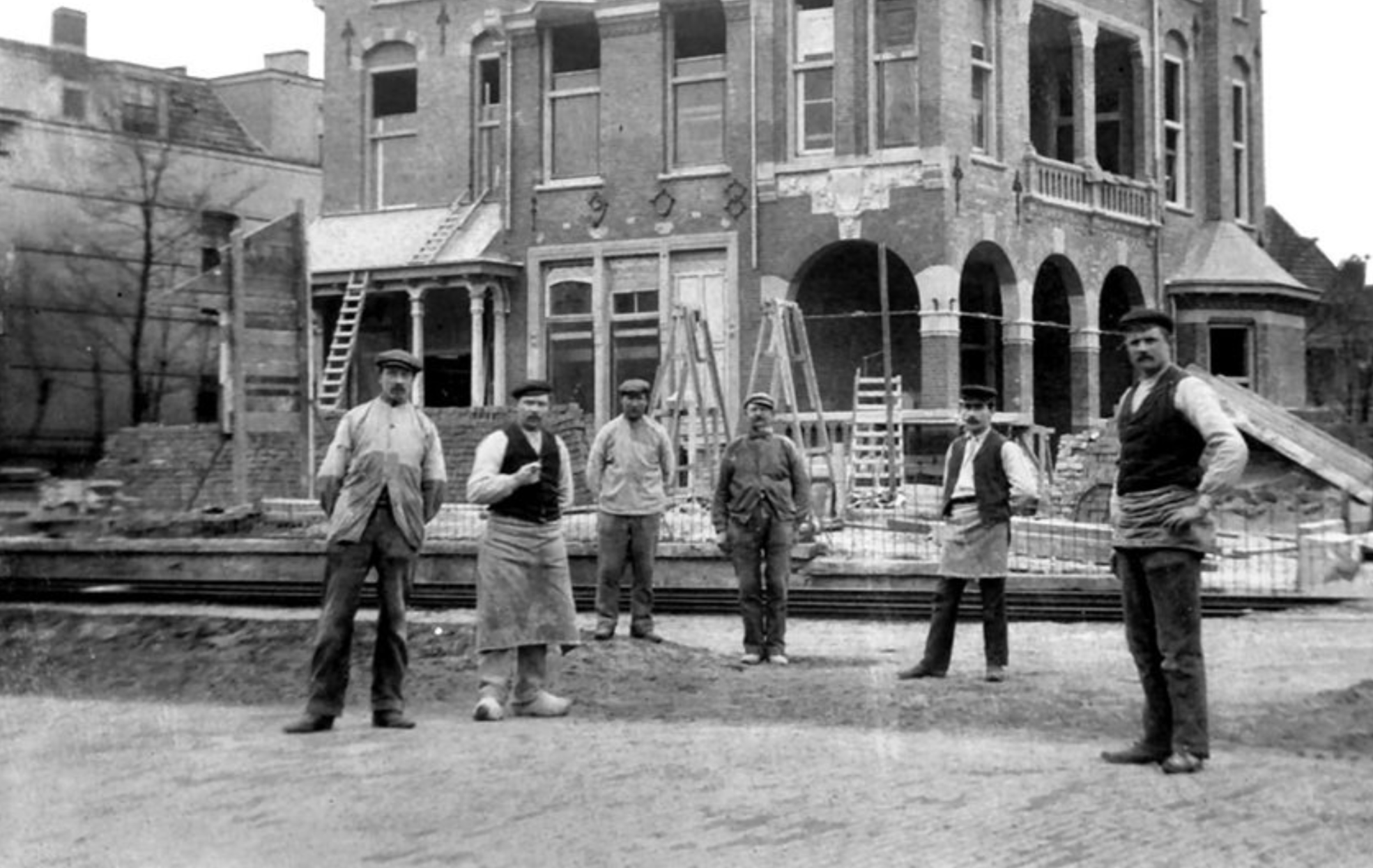 Six construction workers posing in front of a brick building under renovation.