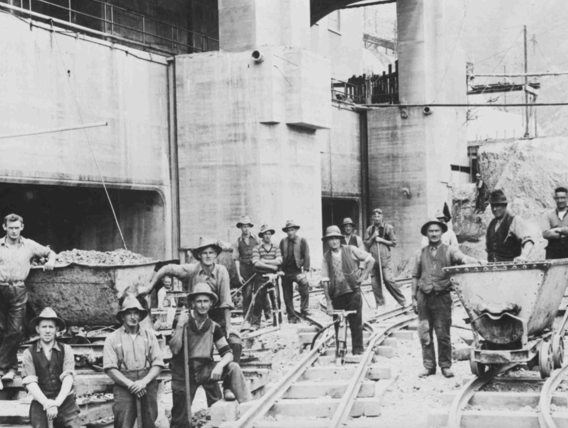 A group of men in hats and work clothes posing in front of a large concrete construction site with mine carts on narrow-gauge tracks.
