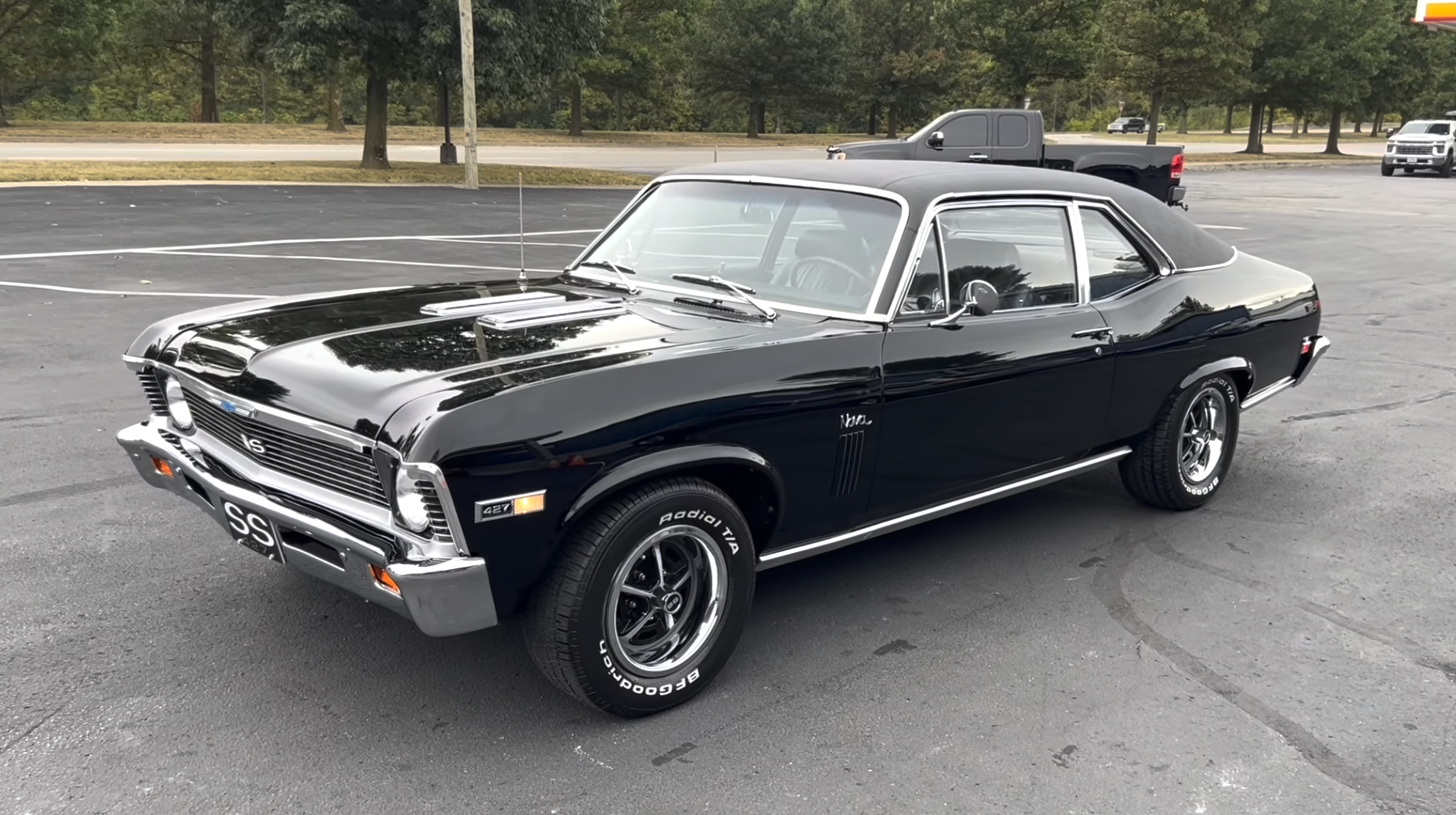 A black classic Chevrolet Nova SS car is parked in a nearly empty lot, surrounded by trees and a few vehicles in the background.