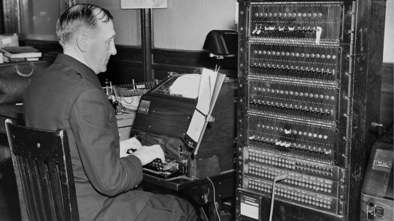 A man in a suit sits at an early computer or switchboard, typing on a keyboard, with a large panel of switches and lights beside him in a vintage office setting.