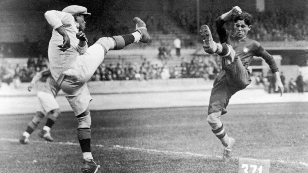 Two soccer players in action, each with one leg raised high, competing for the ball during a match in a crowded stadium. The image is in black and white, capturing an intense moment.