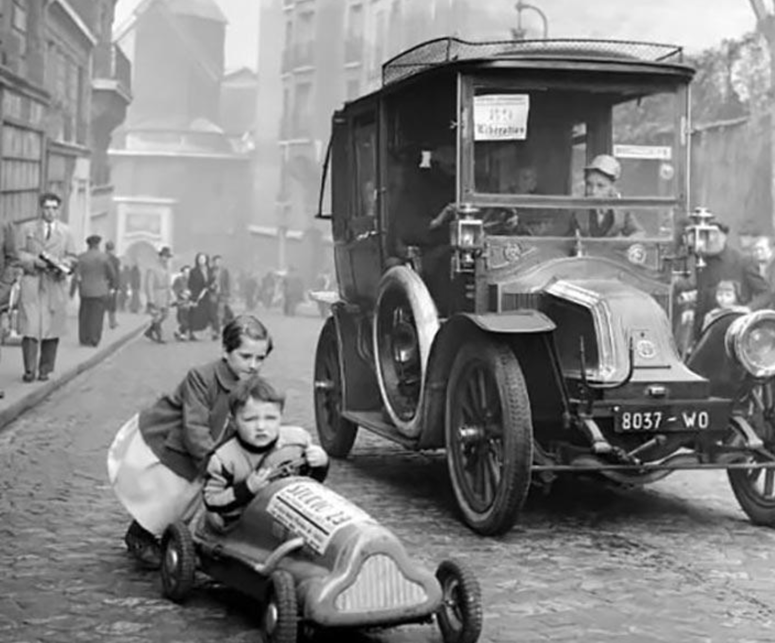 A young girl pushes a small boy in a toy pedal car on a cobblestone street alongside a vintage automobile.