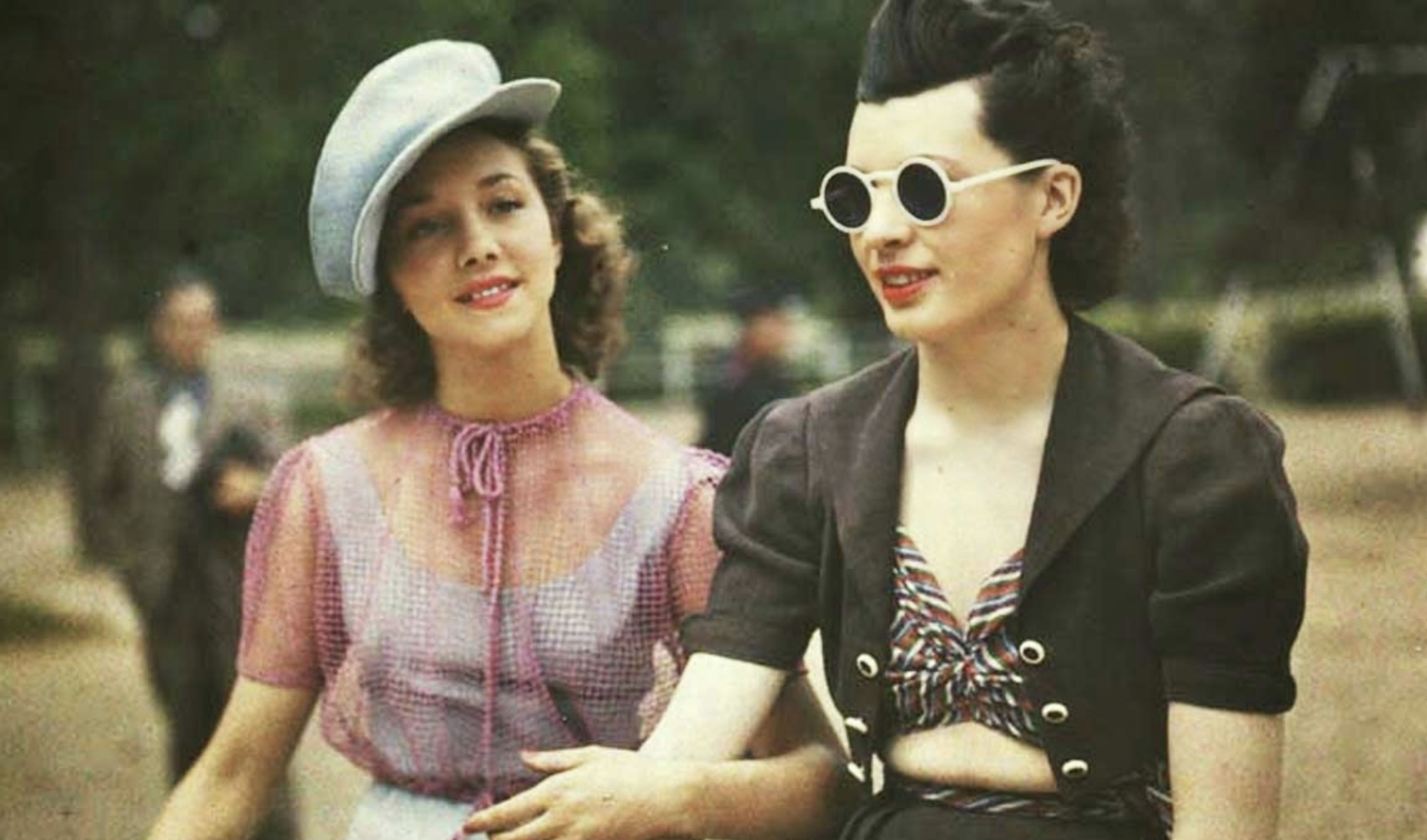 Two young women in 1940s-style clothing outdoors, with one wearing a light-colored cap and the other wearing dark round sunglasses.