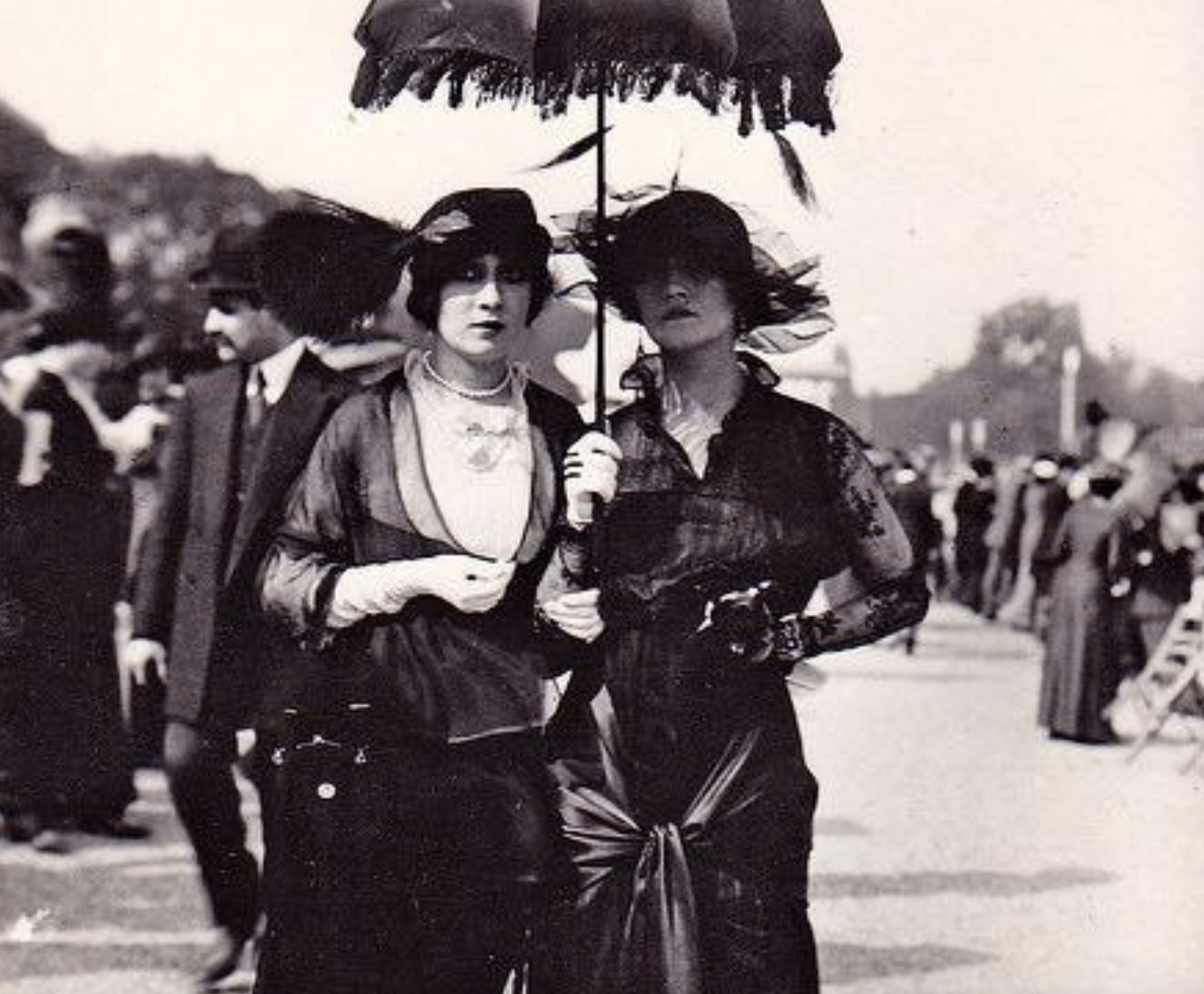 Two women in dark dresses and hats walk outdoors, one holding a fringed parasol.