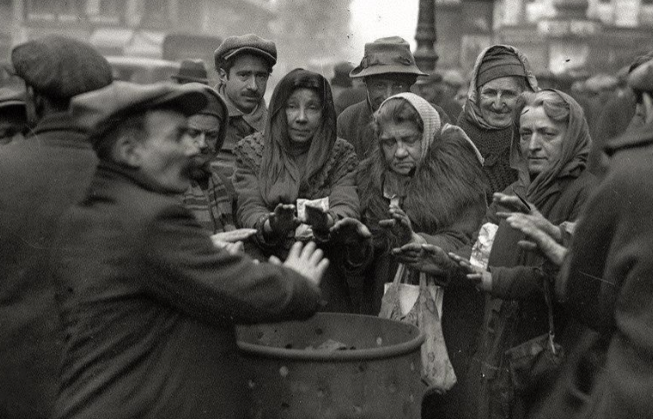 A group of people in winter clothing gather around a street fire barrel, warming their hands.
