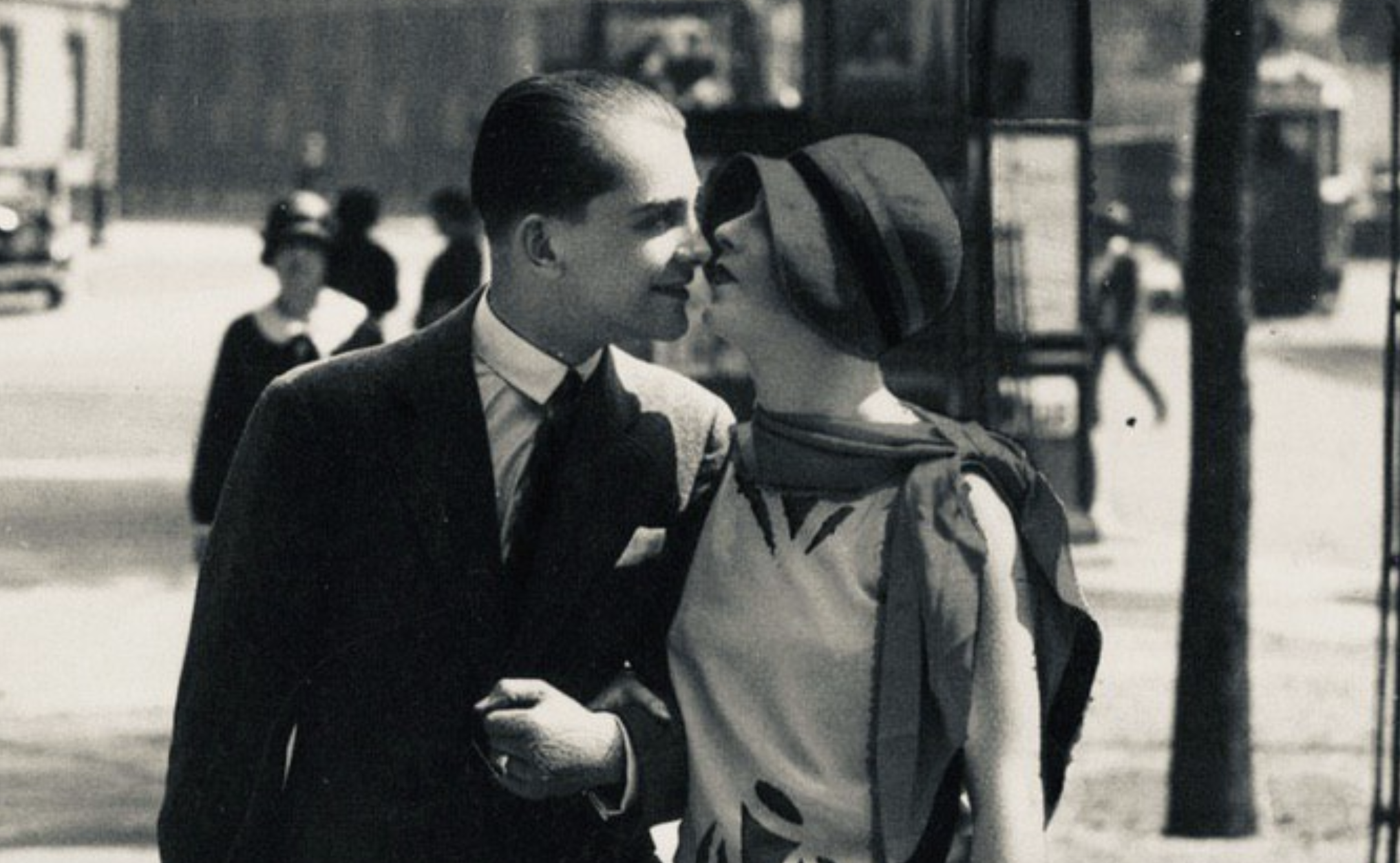 A man in a suit and a woman in a cloche hat lean toward each other as if about to kiss while walking on a city street.