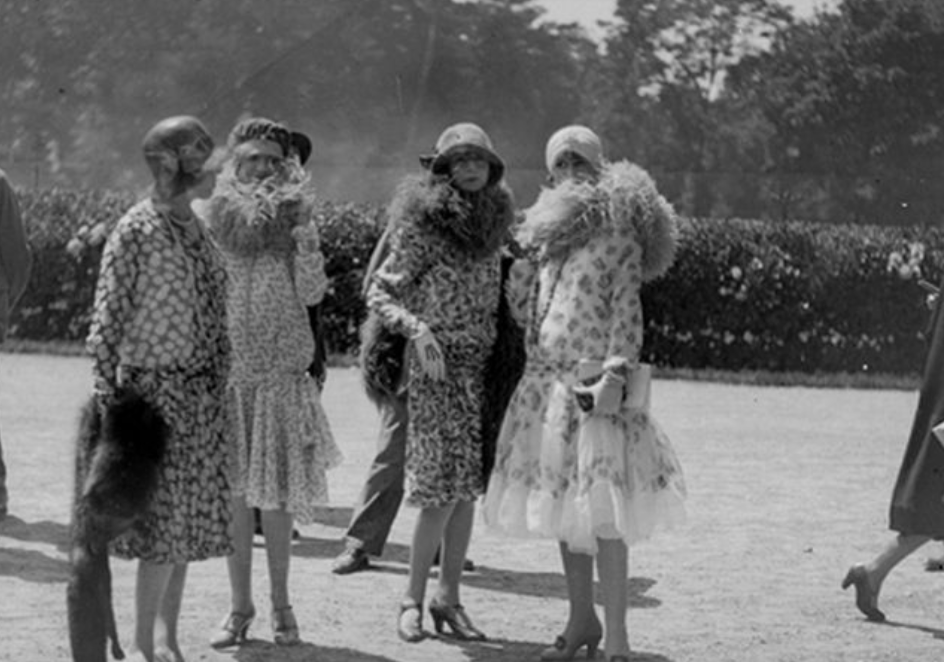 Four women in 1920s fashion, including floral dresses and large feather boas, stand together outdoors.