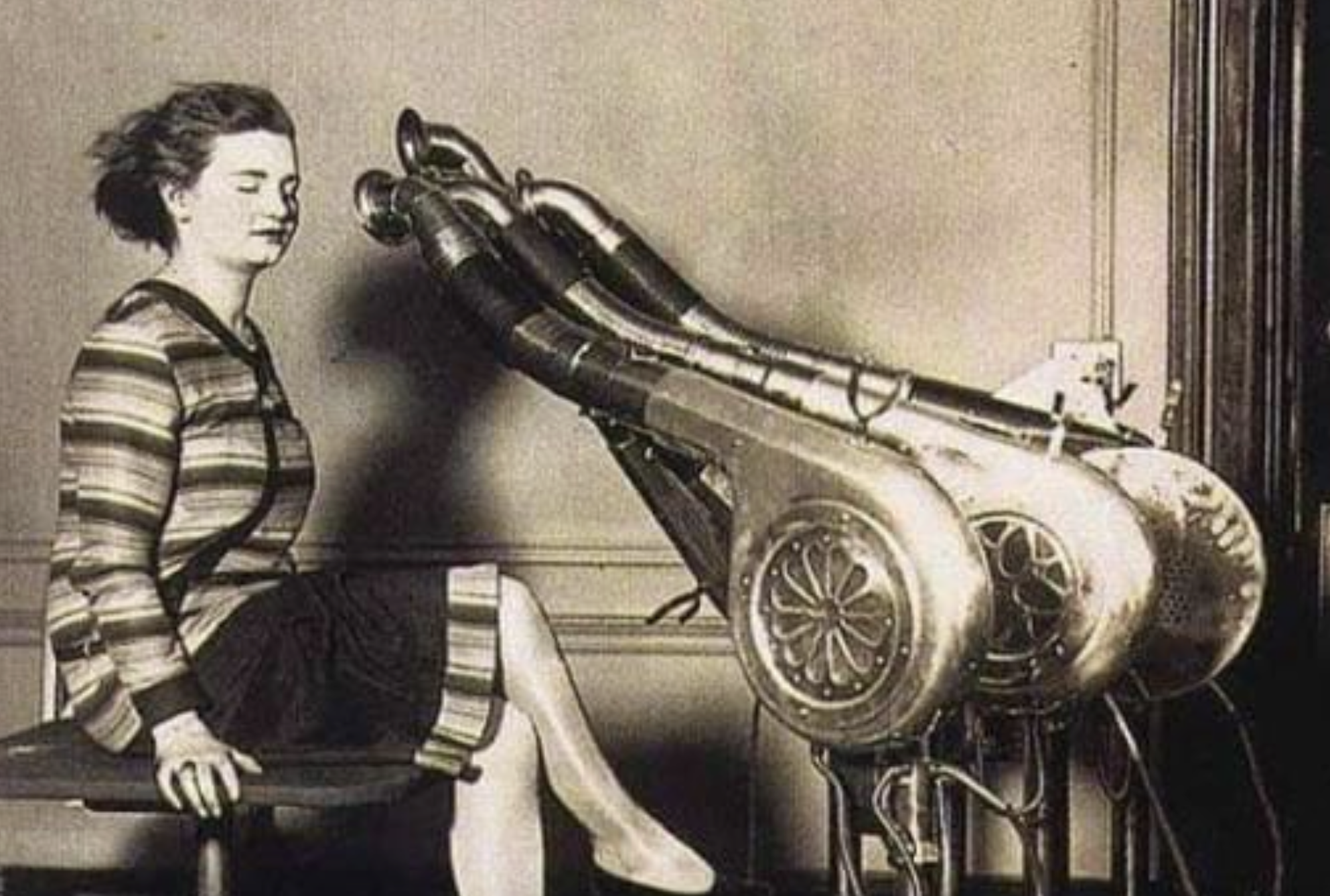 A woman sits with her eyes closed in front of two large, industrial-style hair dryers with multiple nozzles directed toward her head.