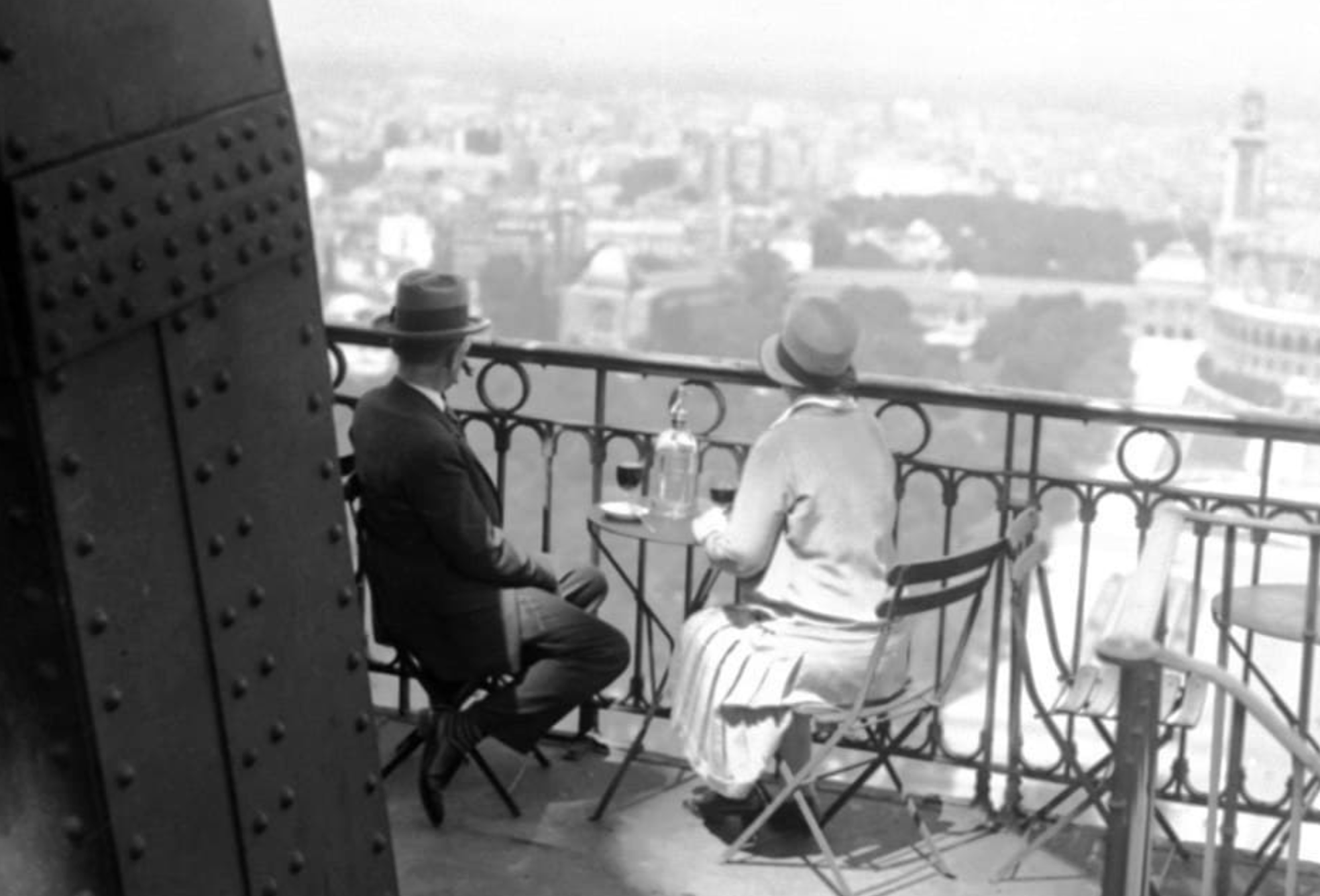 A man and woman in hats sit at a small table on an observation deck overlooking a city.