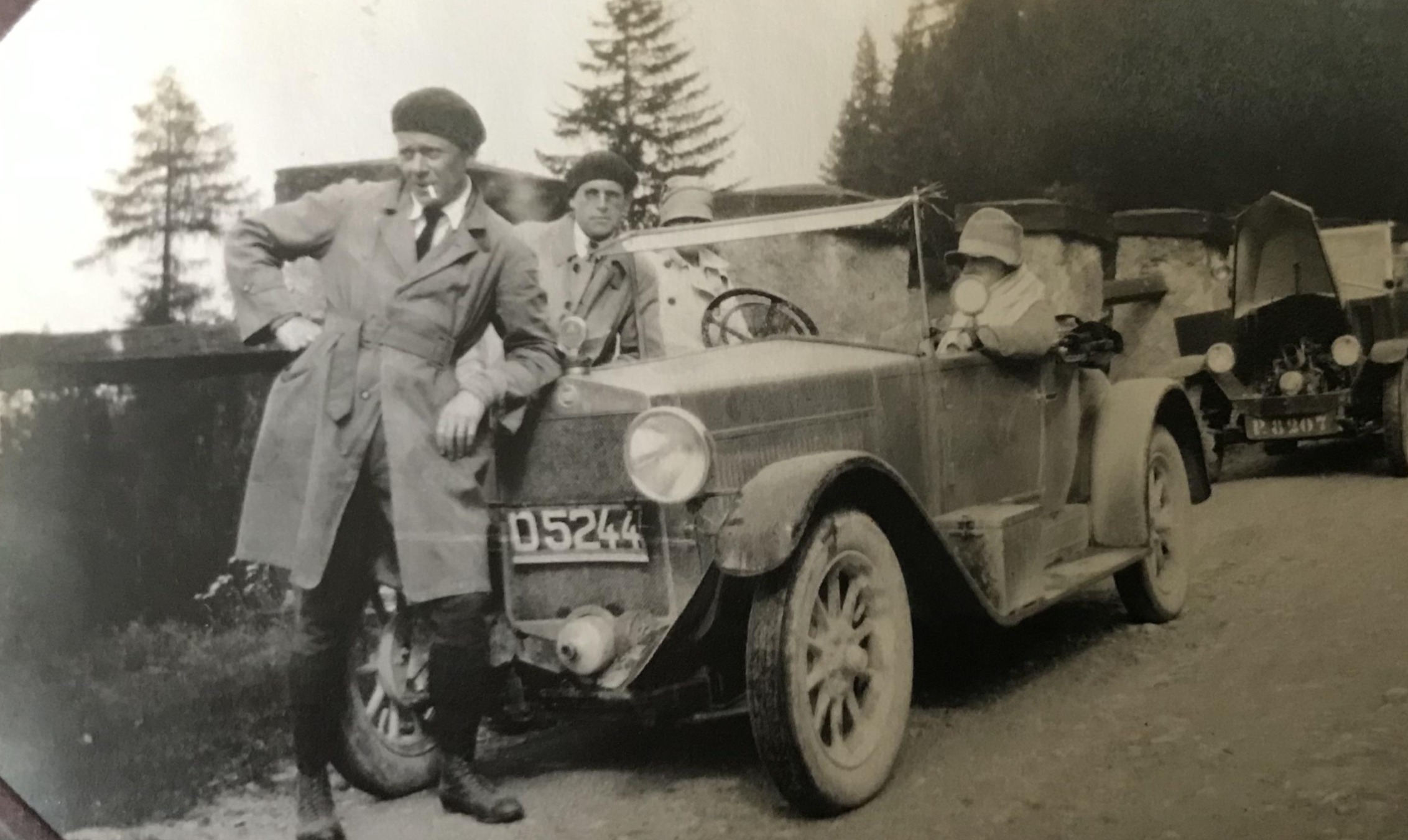 A man in a trench coat and cap stands beside an early 20th-century open-top car with passengers, with a license plate reading D5244.