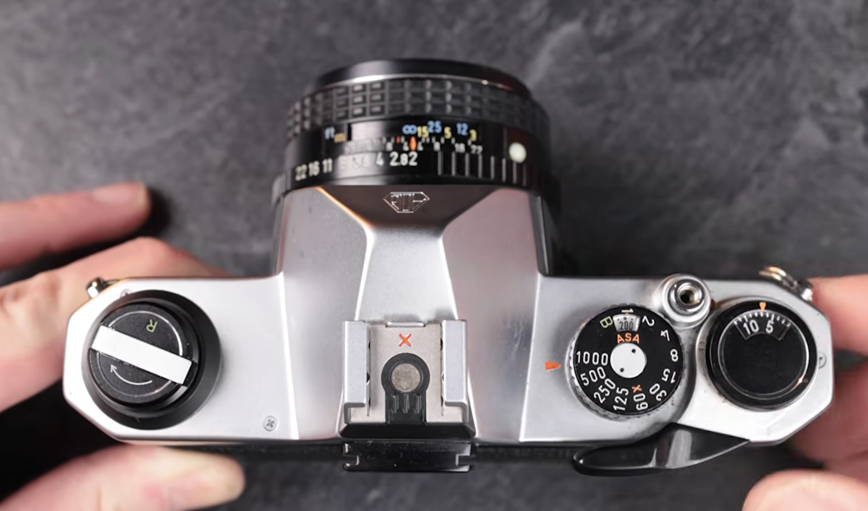 Top-down view of a silver 35mm film SLR camera with a lens attached, held by two hands.