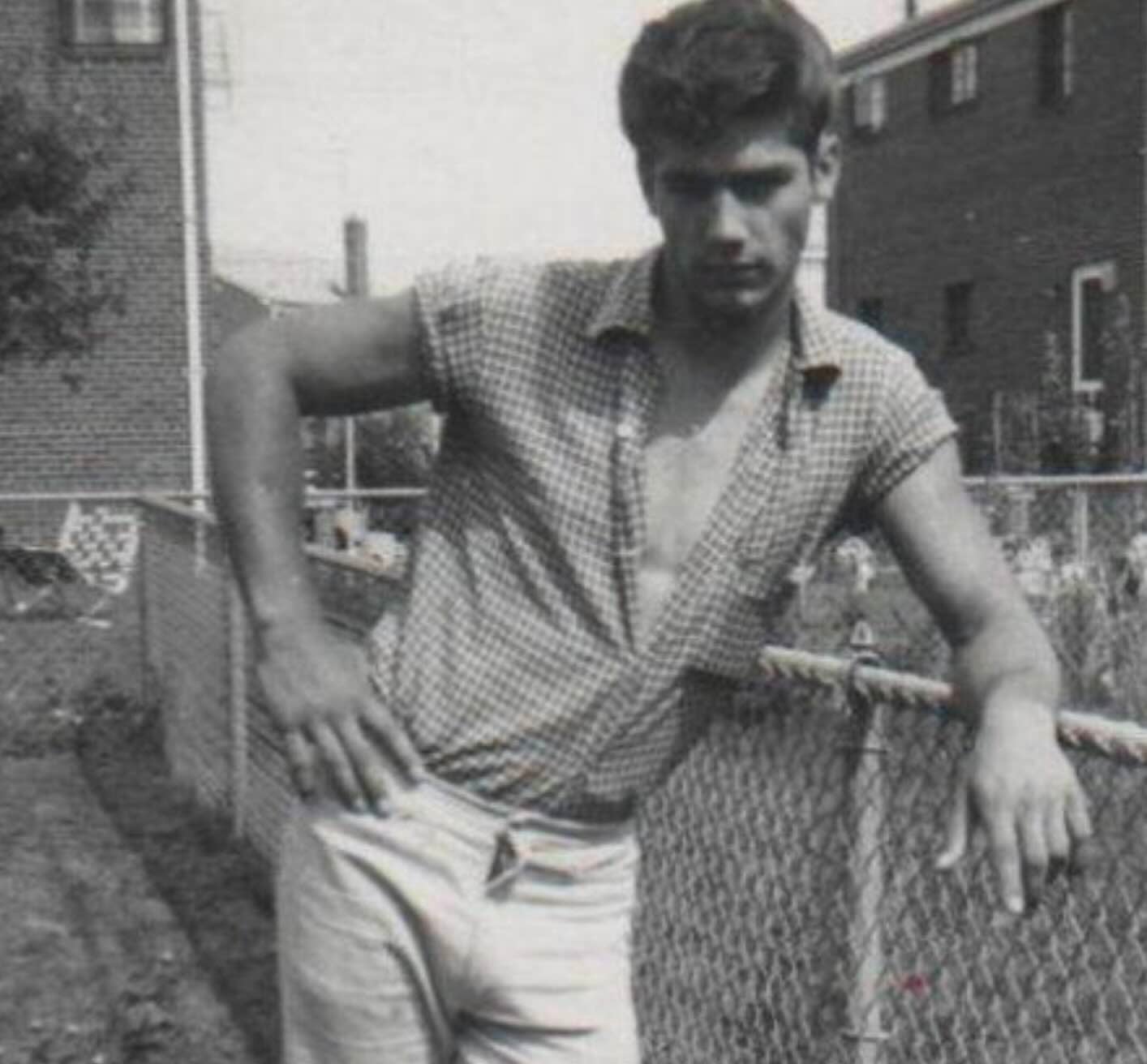A young man in a short-sleeved checkered shirt leaning against a chain-link fence.