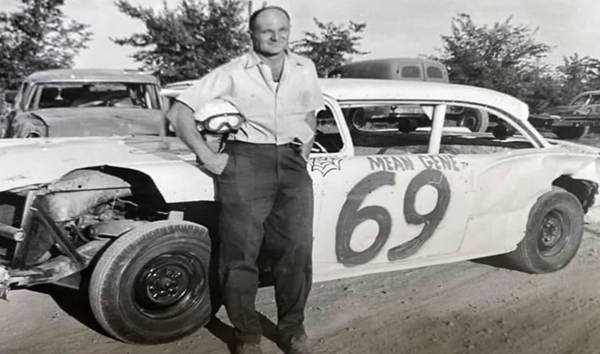 A man wearing a short-sleeved shirt and holding a helmet stands next to a race car numbered 69 with “MEAN GENE” painted on the side.