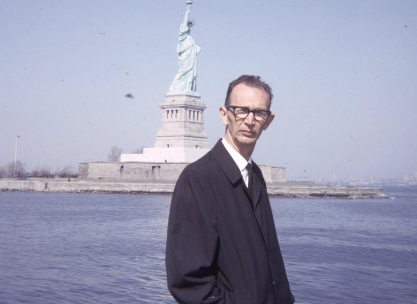 A man in a black coat and glasses stands in front of the Statue of Liberty.
