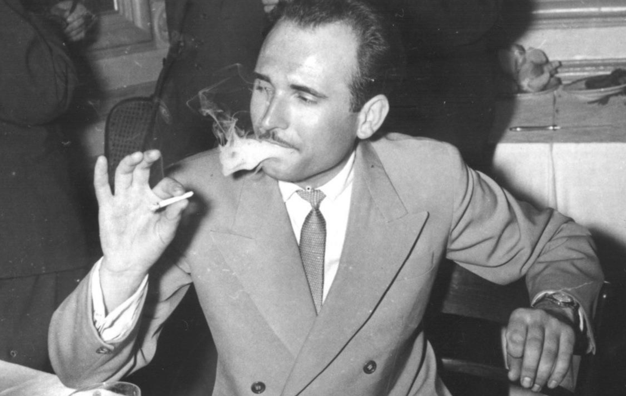 A man in a suit and tie smokes a cigarette while seated at a table.