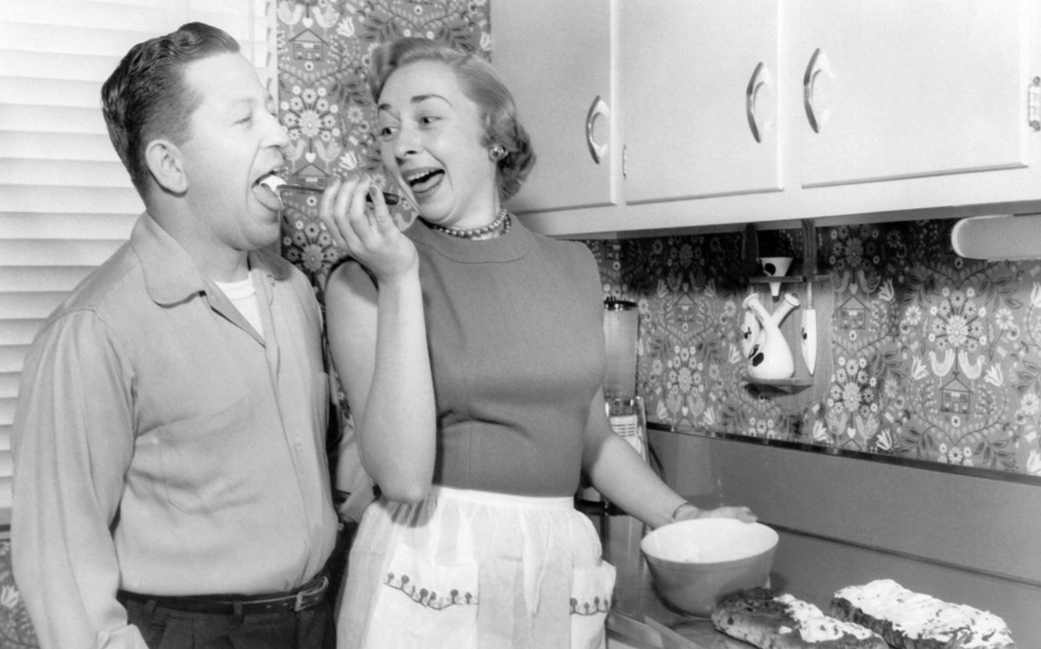 A woman in an apron laughs as she feeds a piece of food to a man in a kitchen.