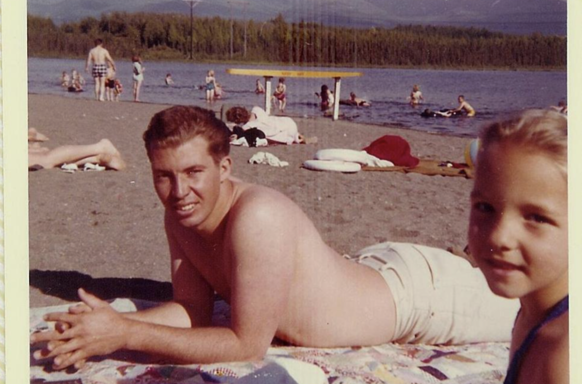 Vintage photo of a young man and child lying on a blanket at a busy beach. A lake, trees, and mountains are visible in the background. Other beachgoers swim and relax nearby.