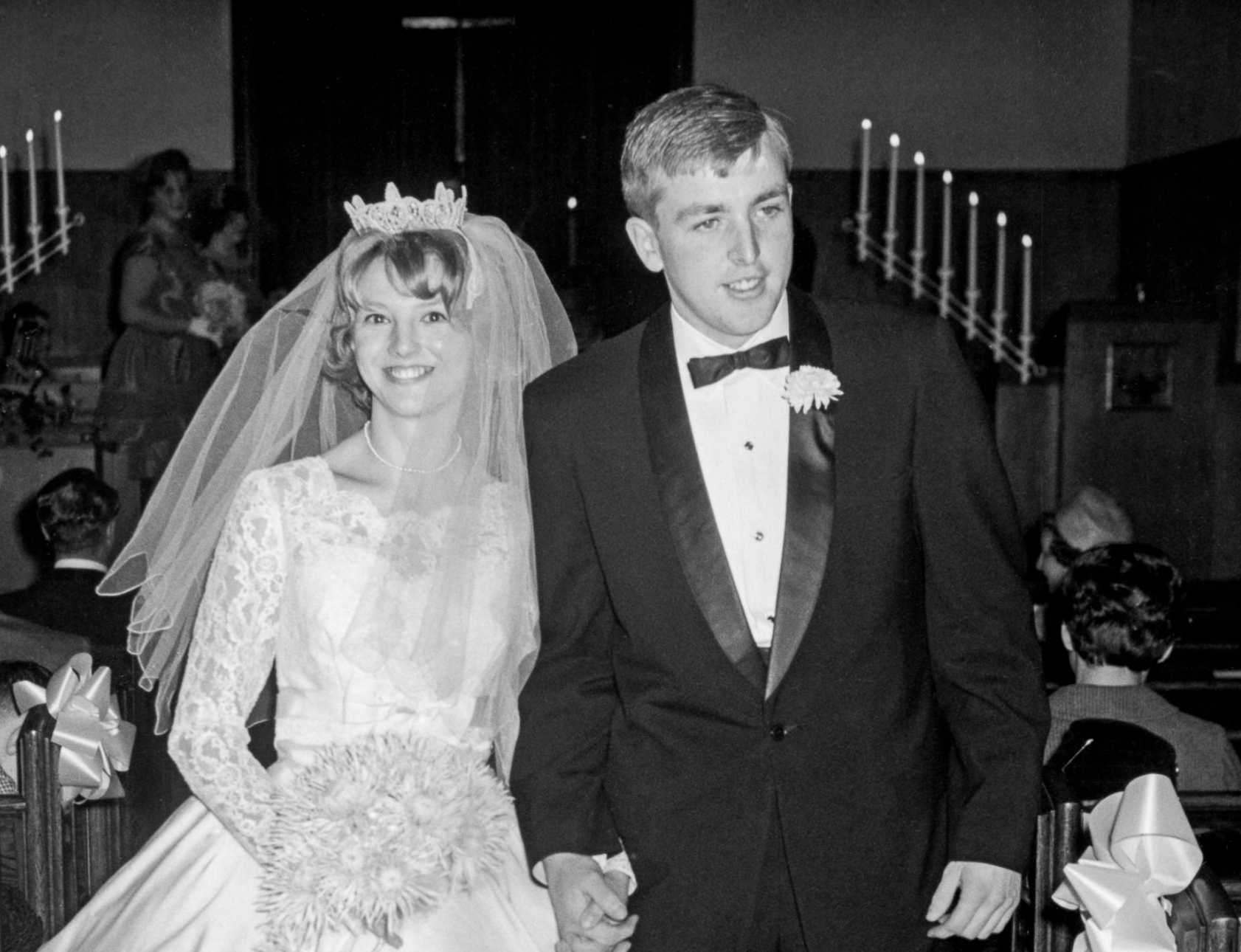 A bride in a lace wedding dress and tiara, and a groom in a tuxedo, walk hand-in-hand down an aisle.