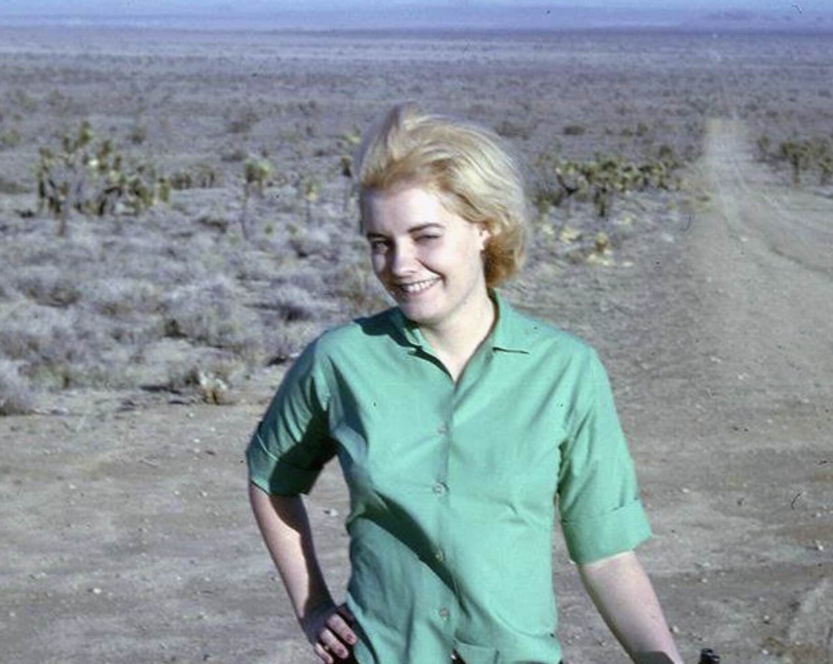 A smiling woman with blonde hair wearing a green button-down shirt stands in a desert landscape.
