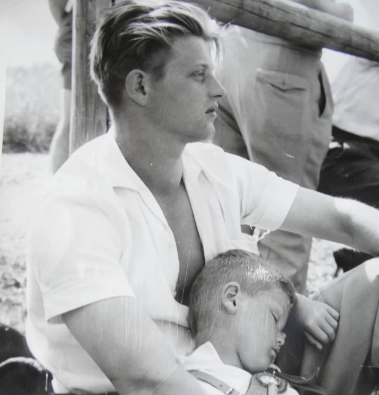 A black and white photograph of a young man sitting outdoors with a small child sleeping in his lap.