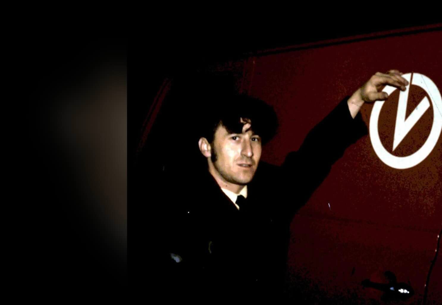 A man in a dark jacket reaching up to touch a white circular logo on a red surface.