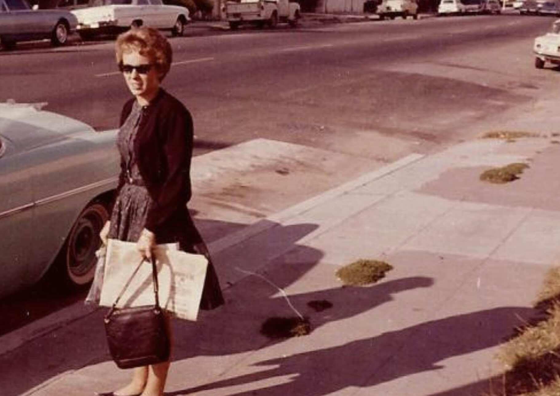 A woman in a dark dress and sunglasses stands on a sidewalk beside a parked car, holding a bag and some papers.
