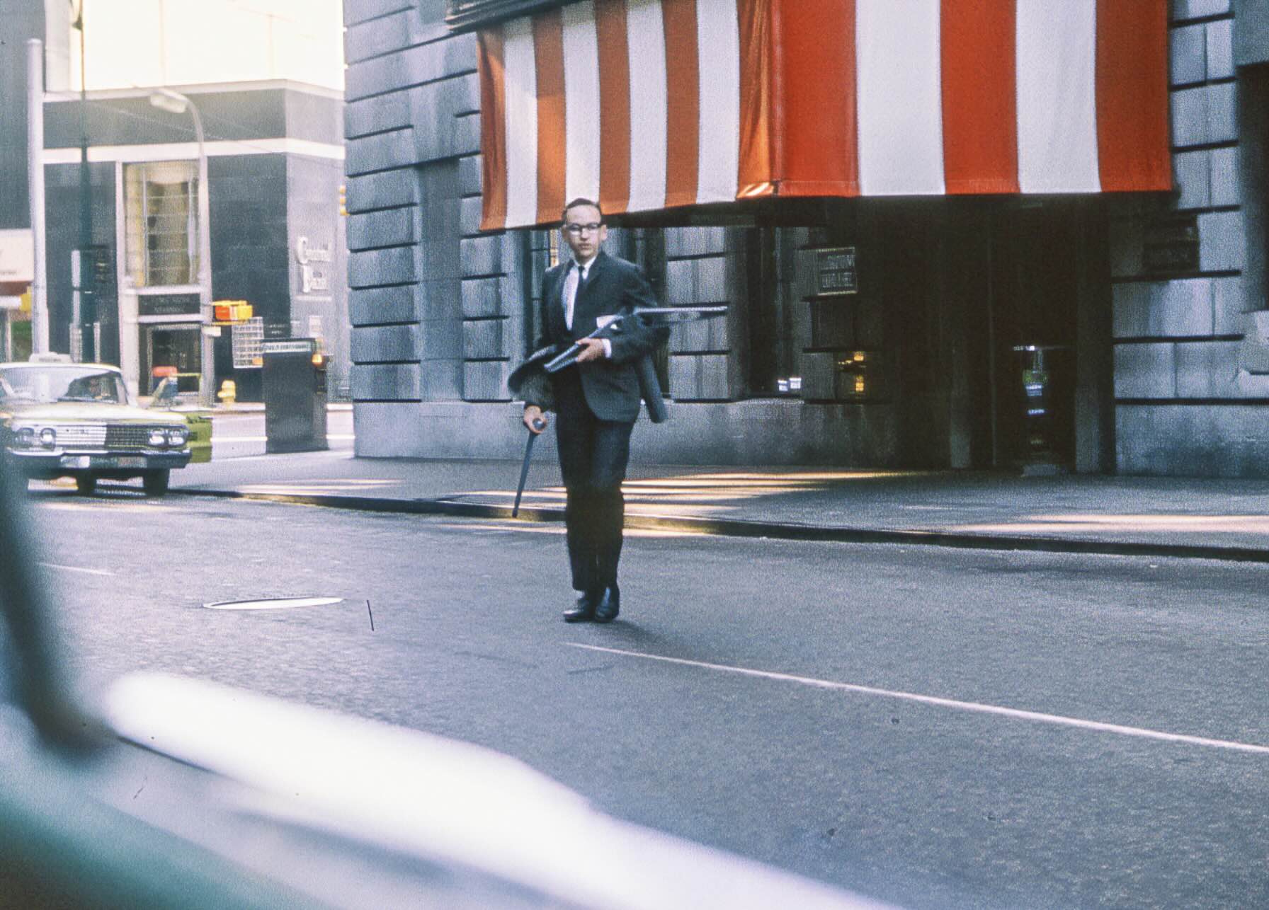 A man in a suit and glasses carries a briefcase and umbrella while walking across a city street.