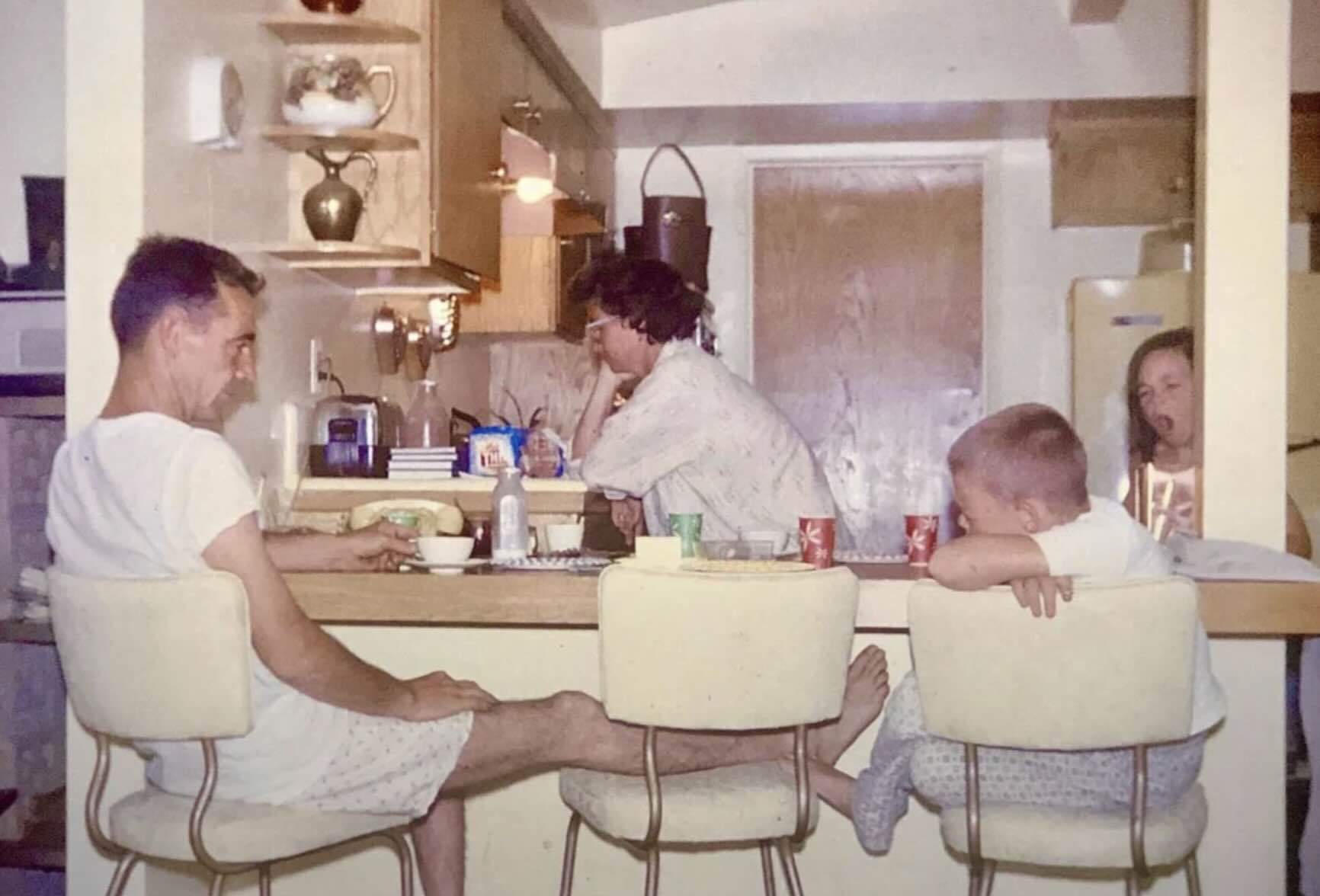 A family sits at a kitchen counter, with a man, woman, and young boy eating while a girl looks on from the background.