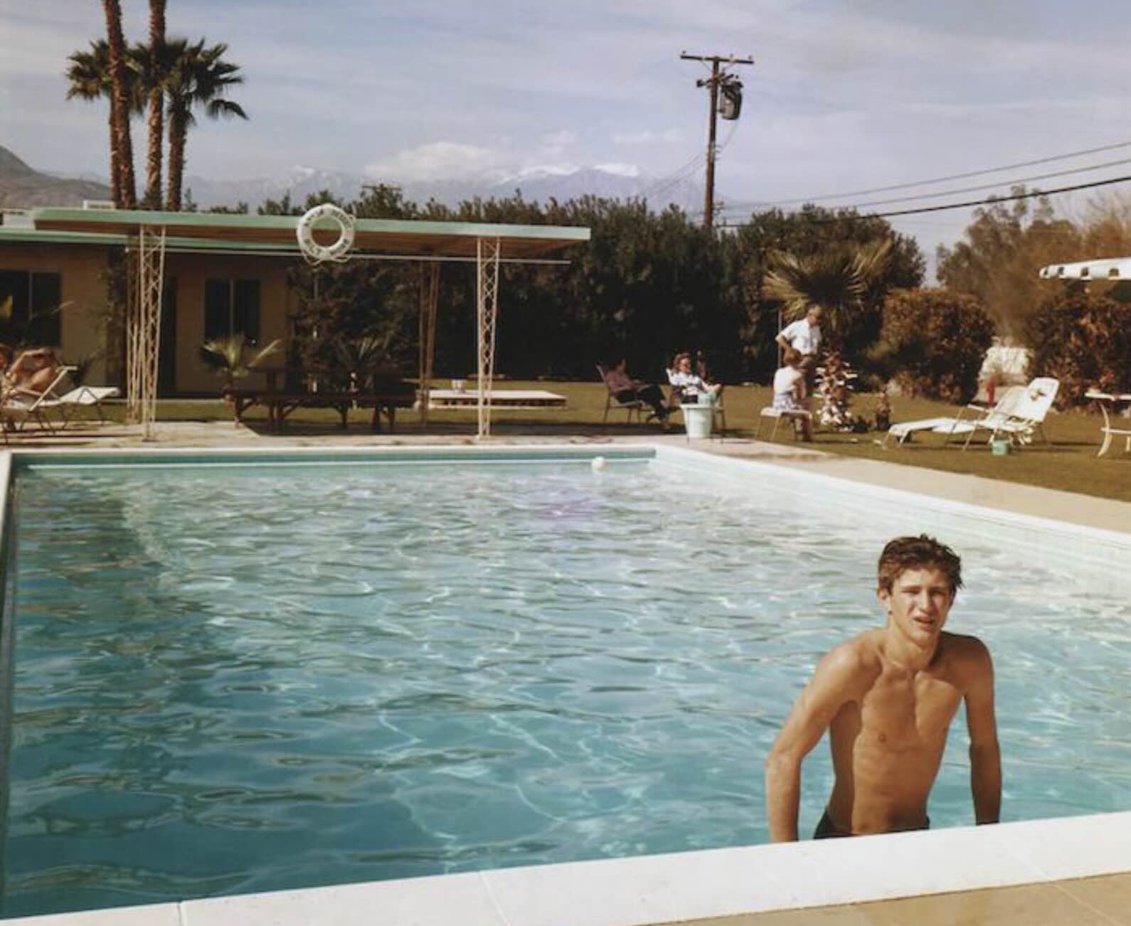 A shirtless young man stands in a swimming pool while people relax on lounge chairs on the lawn behind him.
