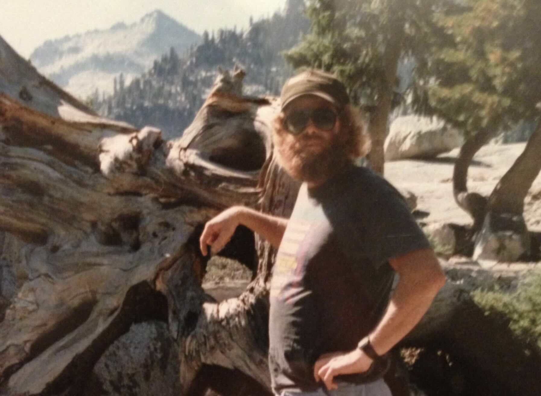 A bearded man wearing a baseball cap and sunglasses stands in front of a large fallen tree in a mountain landscape.