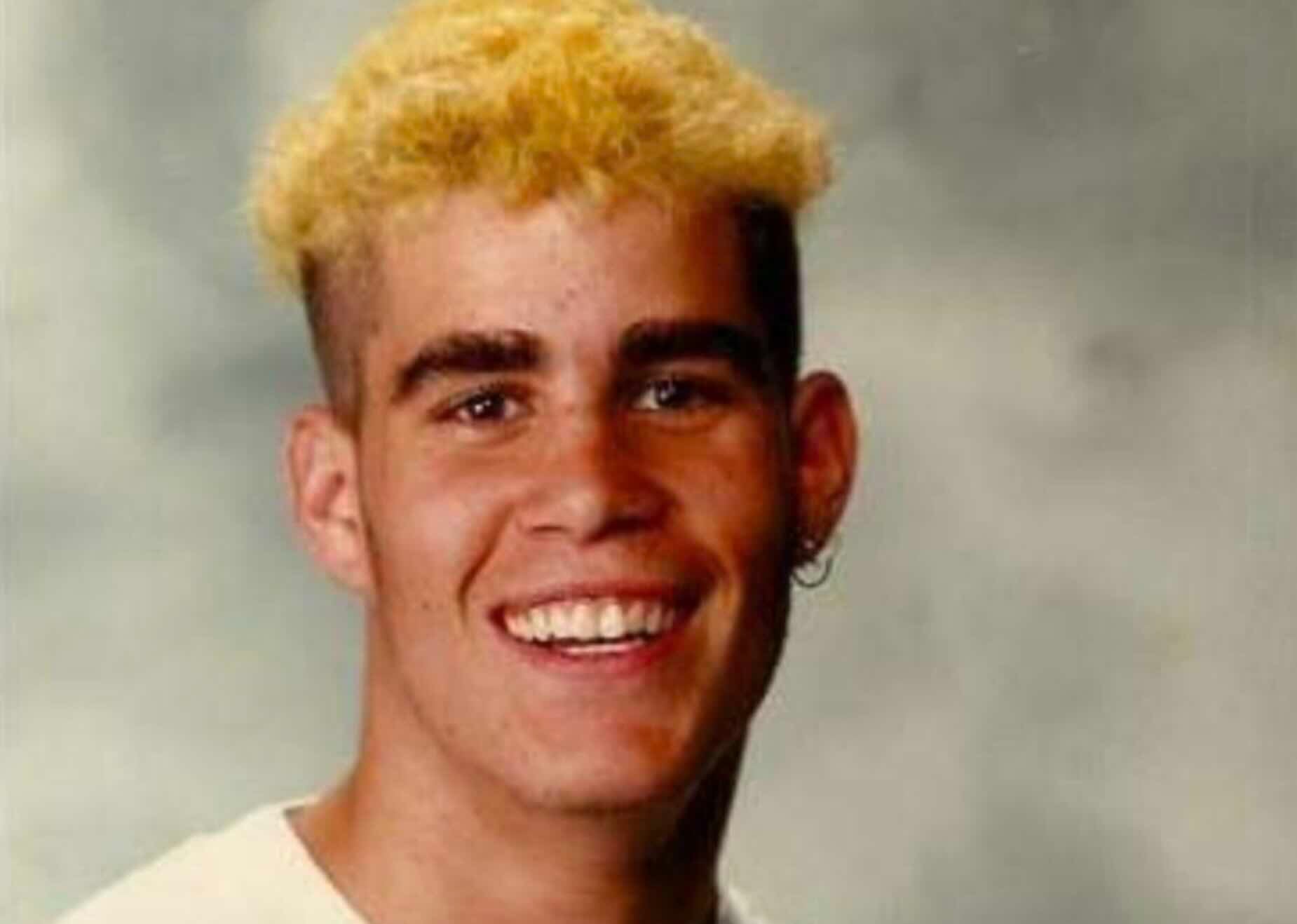 A portrait of a smiling young man with a short, bleached-blonde haircut and a small hoop earring.