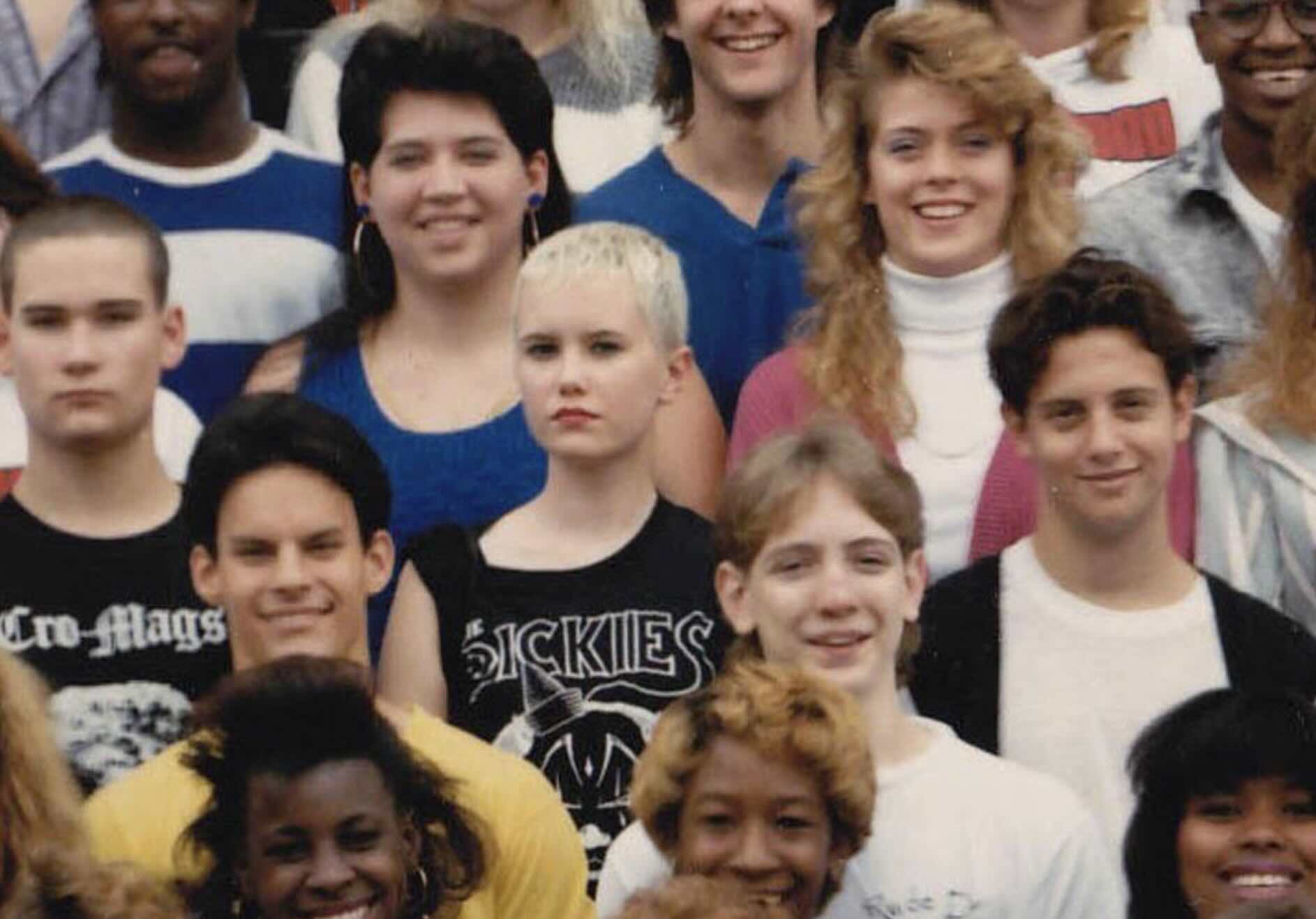 A group of high school students posing for a class photo, including a girl in the center wearing a band t-shirt that says “Sickies.”