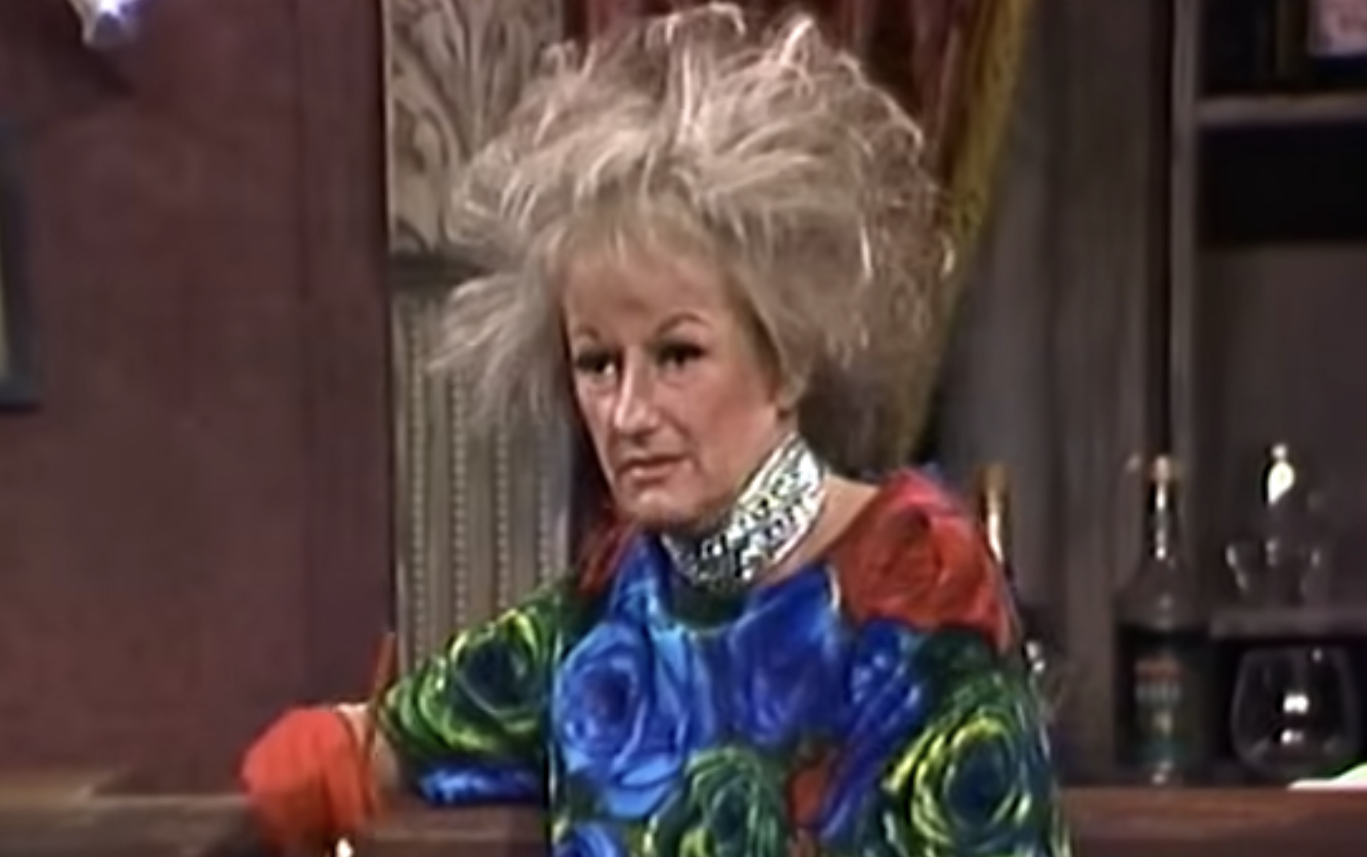 Phyllis Diller with signature wild hair and a blue floral shirt wearing a metallic choker.