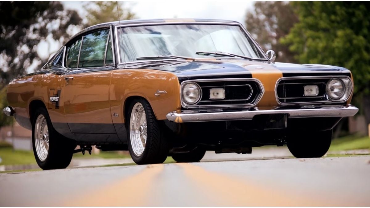A classic two-tone muscle car, black and gold, parked on a street with trees and houses blurred in the background. The vehicle has shiny chrome wheels and a sporty, vintage design.
