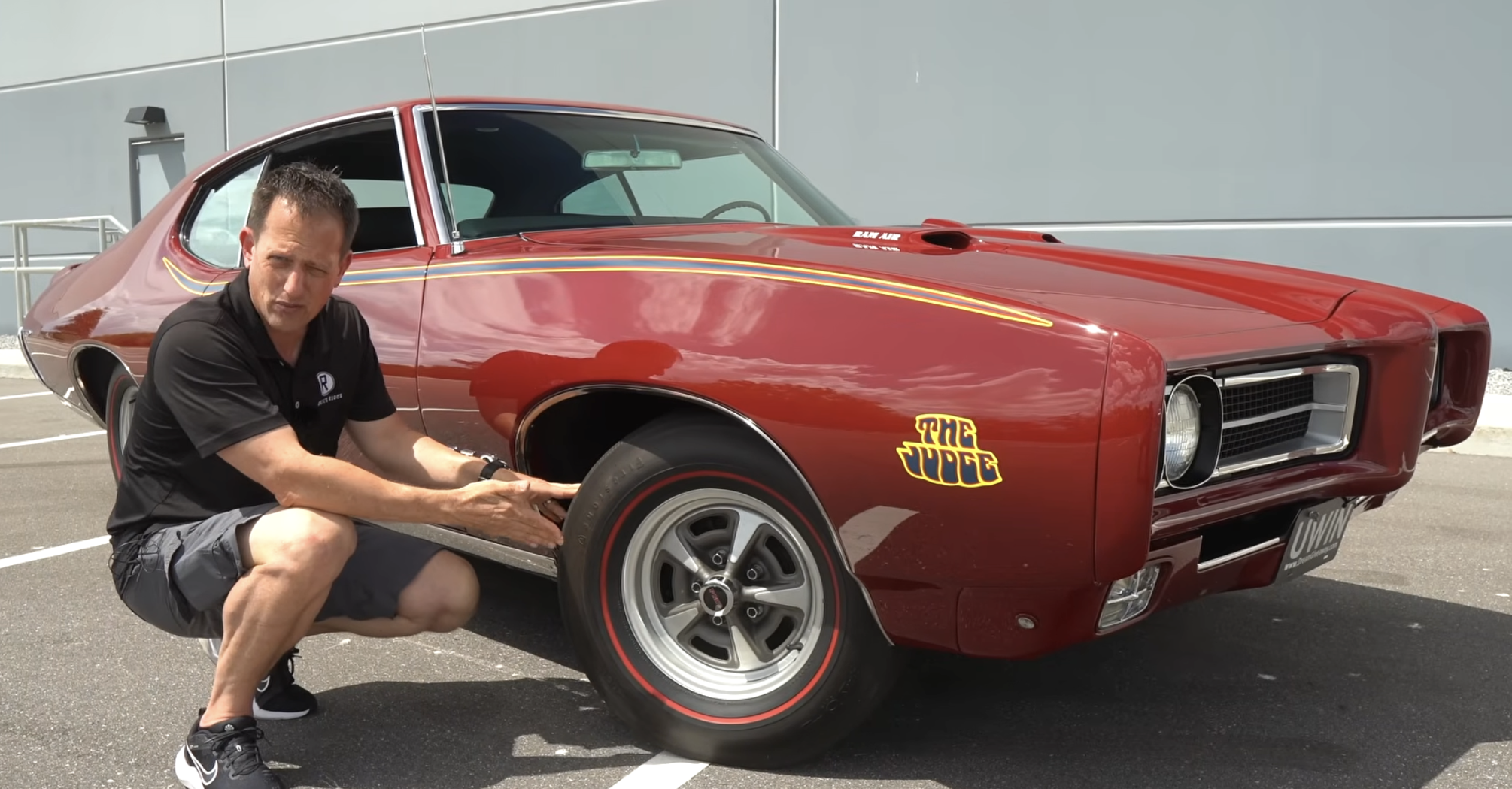 A man crouches beside a red classic muscle car with “The Judge” decal, pointing at the front wheel in a parking lot.