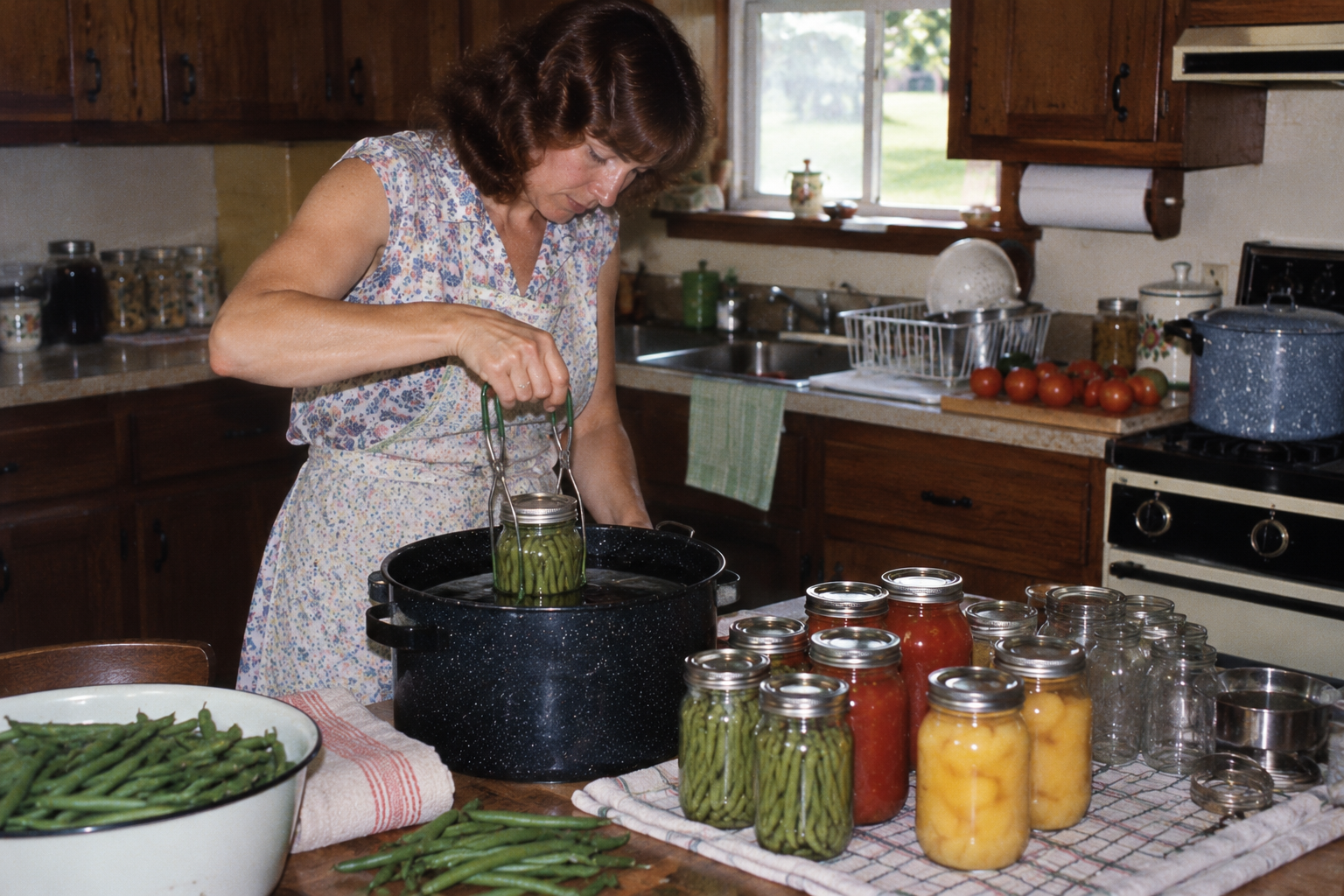 A woman in a floral apron uses jar lifter tongs to lower a glass jar into a large pot of water on a kitchen counter filled with other jars of preserved vegetables and fruit.