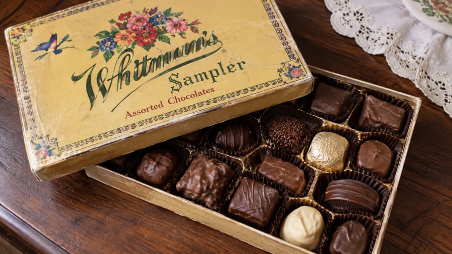 An open Whitman’s Sampler box of assorted chocolates sits on a wooden table next to a lace doily. The box features floral designs and gold and brown wrapped chocolates are visible inside.